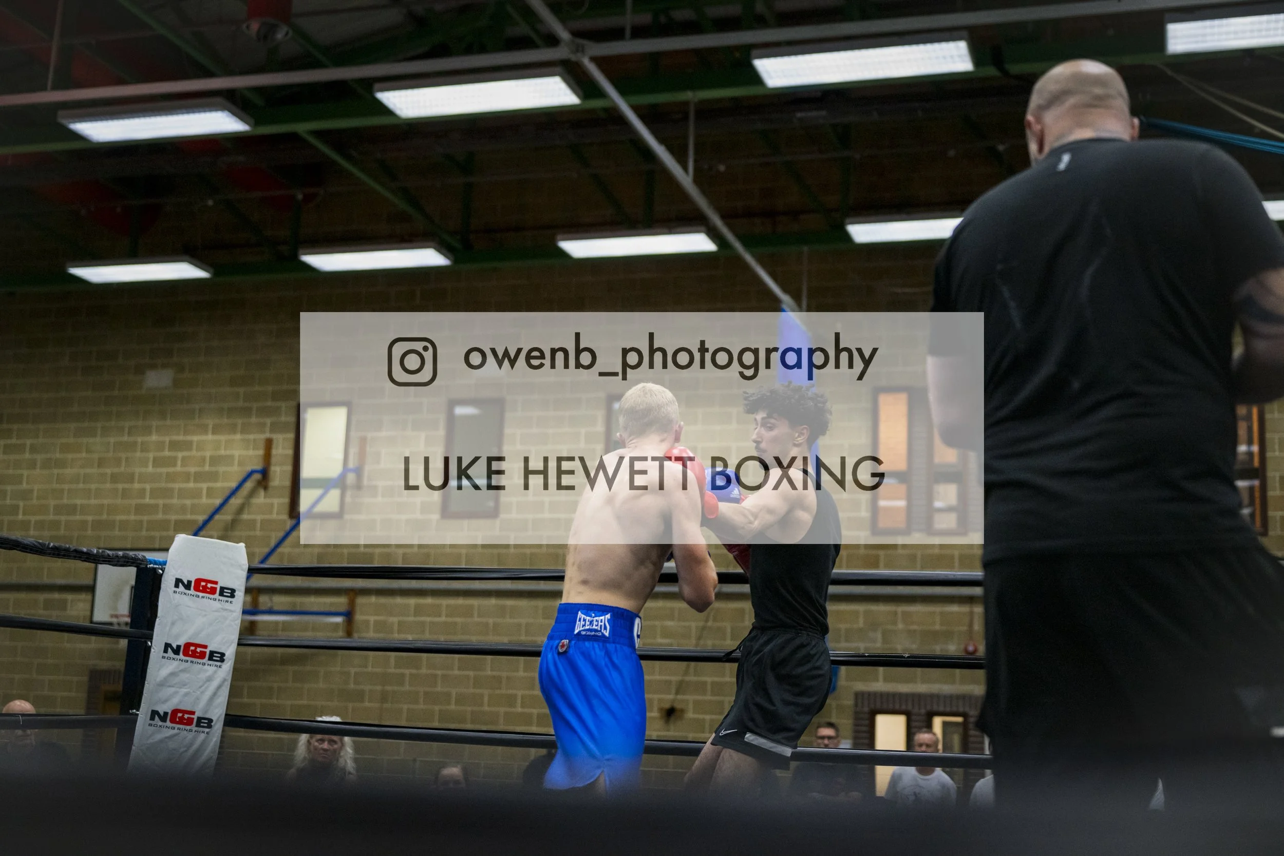 Two young male boxers in boxing gloves and shorts sparring in a boxing ring, with a referee observing nearby, inside a gym with brick walls and fluorescent lighting.