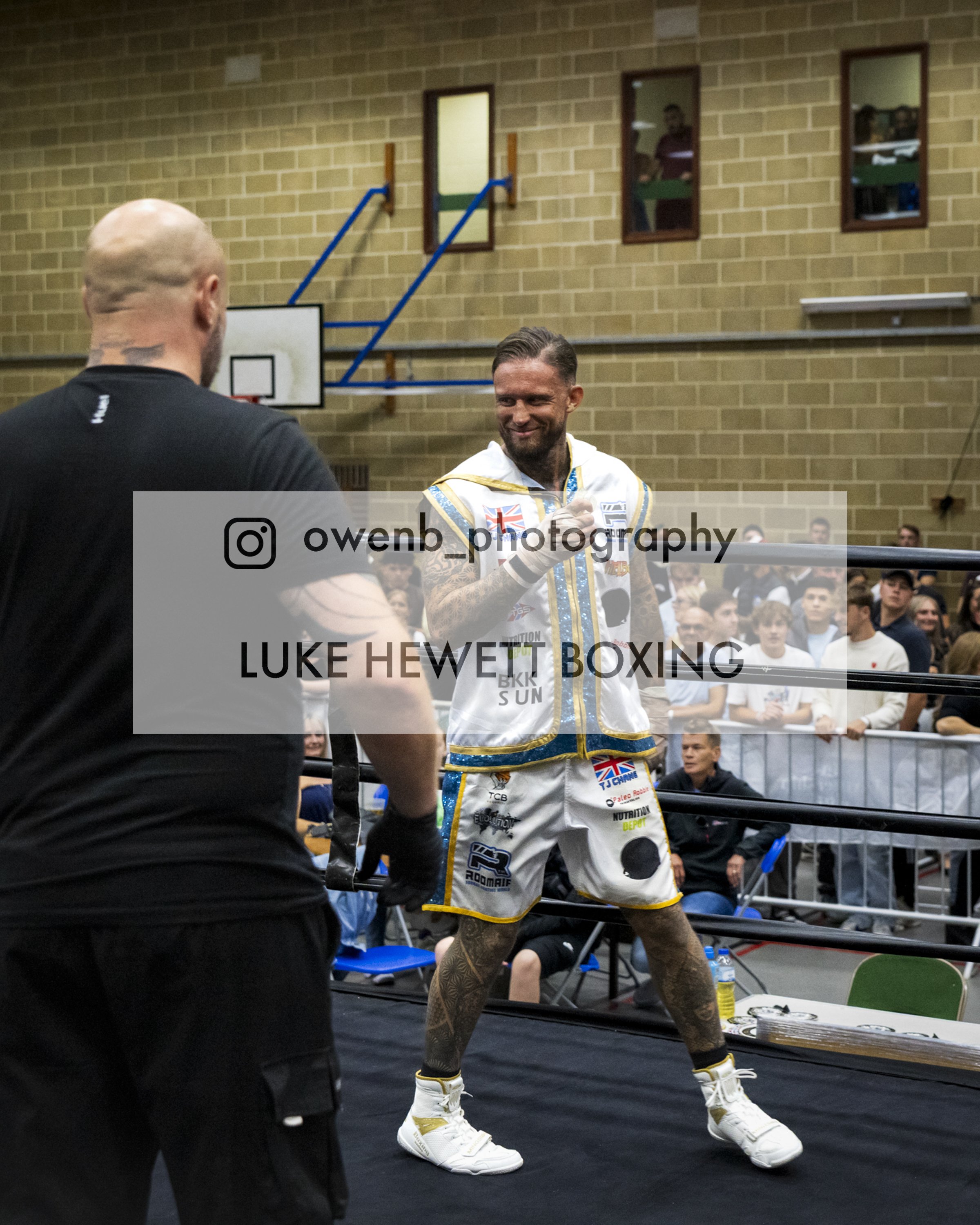 A boxer in boxing gear, including a white and gold robe, standing in a ring, smiling and leaning on the ropes. Opposite him is a bald man with tattoos on his neck, facing away from the camera. In the background, an audience sits and watches the match