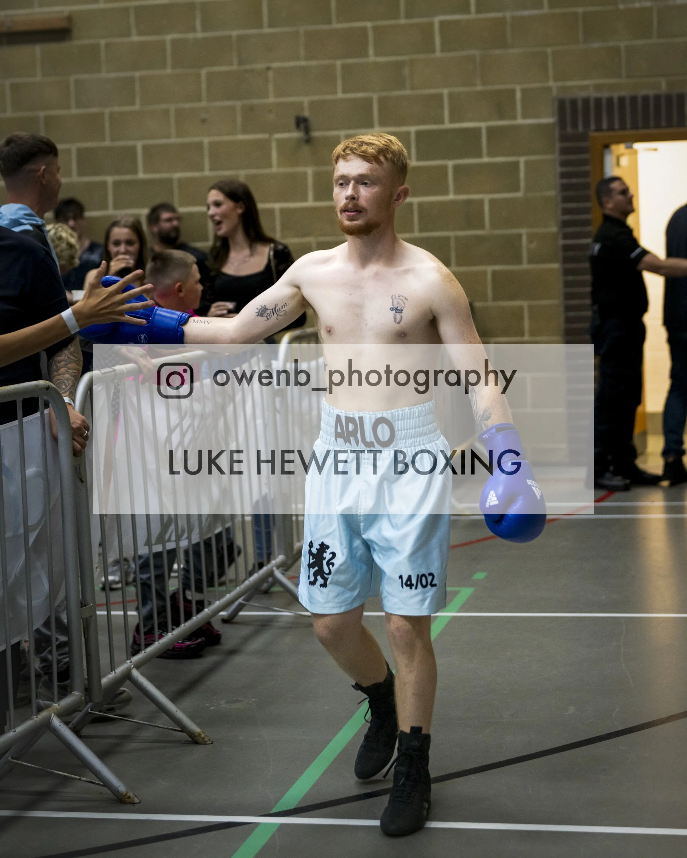 A shirtless male boxer with red hair and tattoos on his chest and arm, wearing white boxing shorts with black print, black boxing shoes, stands in a gymnasium next to a metal fence, holding blue boxing gloves, shaking hands with a person. In the back