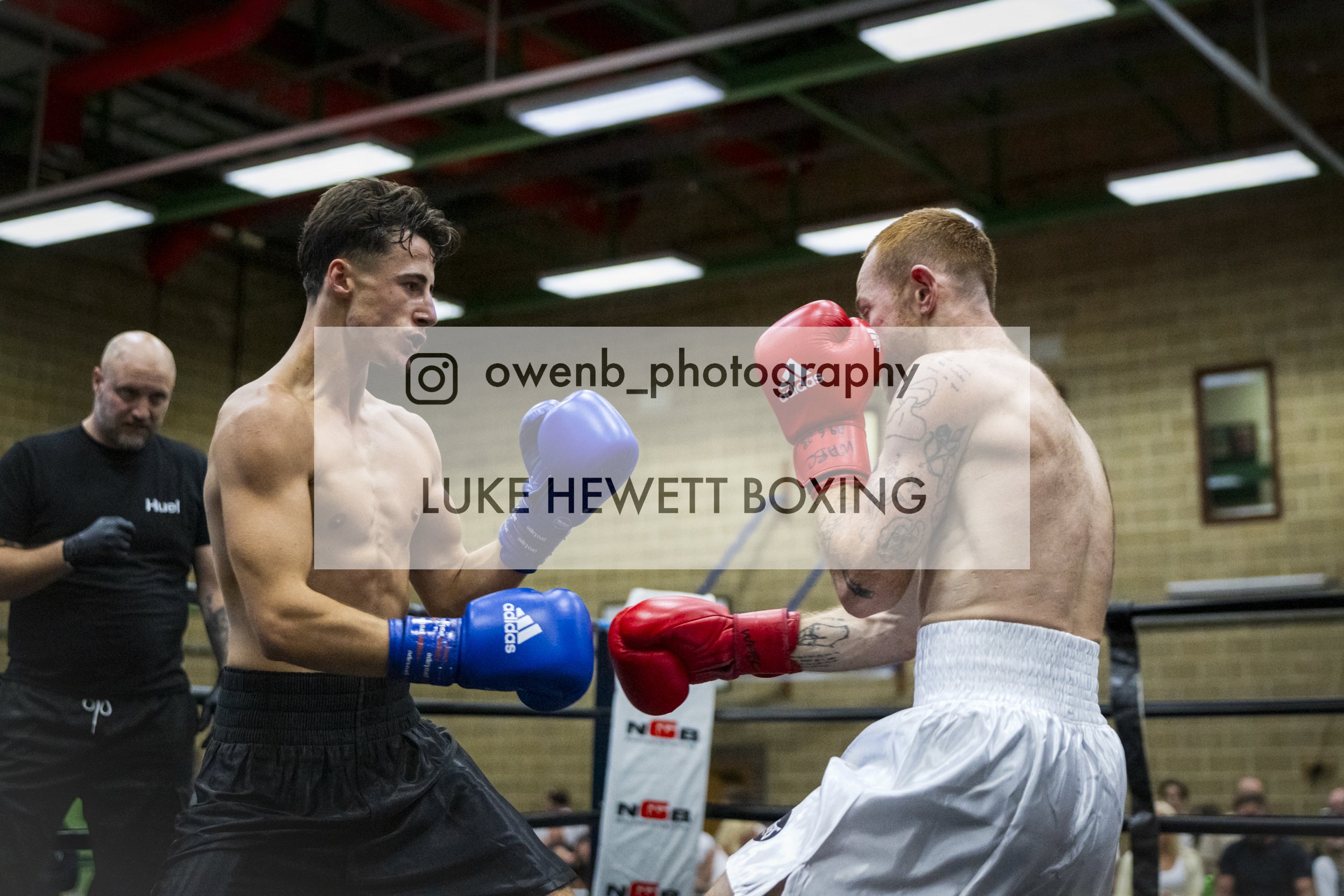 Two shirtless male boxers in a ring, facing each other with fists raised, wearing boxing gloves; referee in black shirt in the background; indoor gym with wooden walls and ceiling lights.