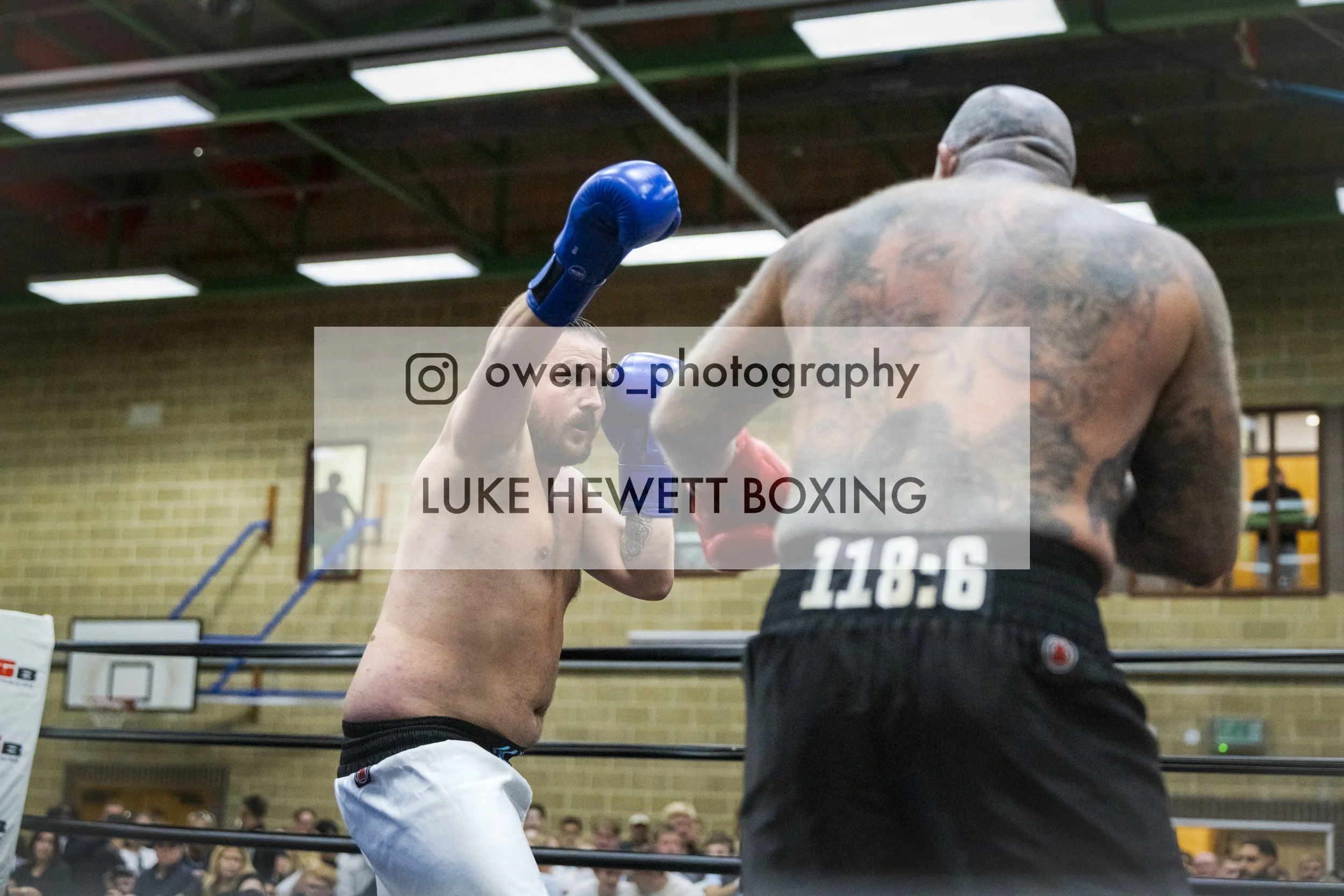 A boxing match in a gym with one fighter throwing a punch at his opponent. The fighter is shirtless, wearing white shorts, and blue gloves. The opponent has tattoos on his back, is wearing black shorts, and has grey hair.