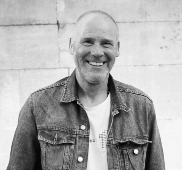 Black and white photo of smiling middle-aged man with a shaved head wearing a denim jacket over a t-shirt, standing against a brick wall.