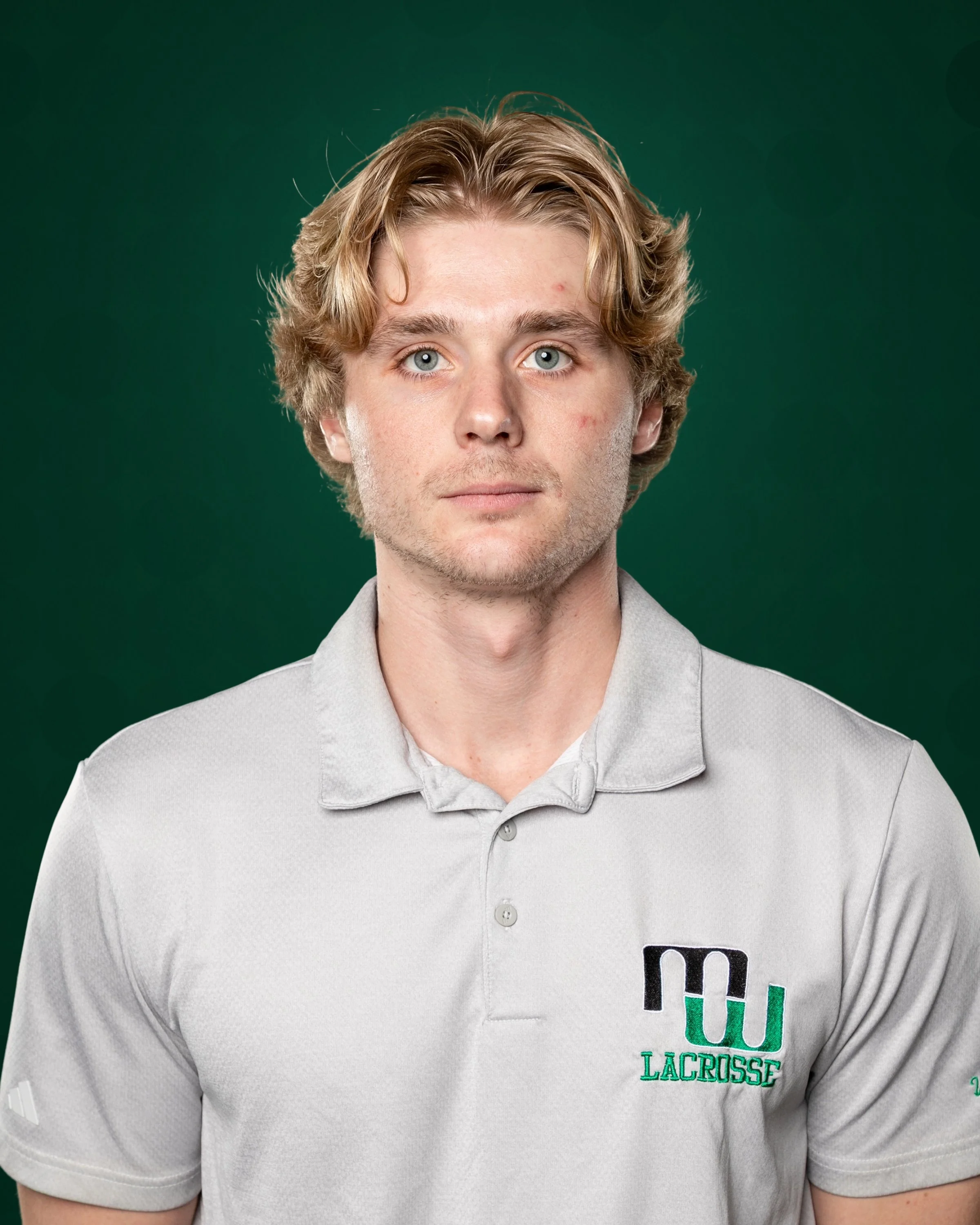 A young man with blonde hair standing on a sports field at night, wearing a black jacket with a lacrosse logo, with floodlights in the background.