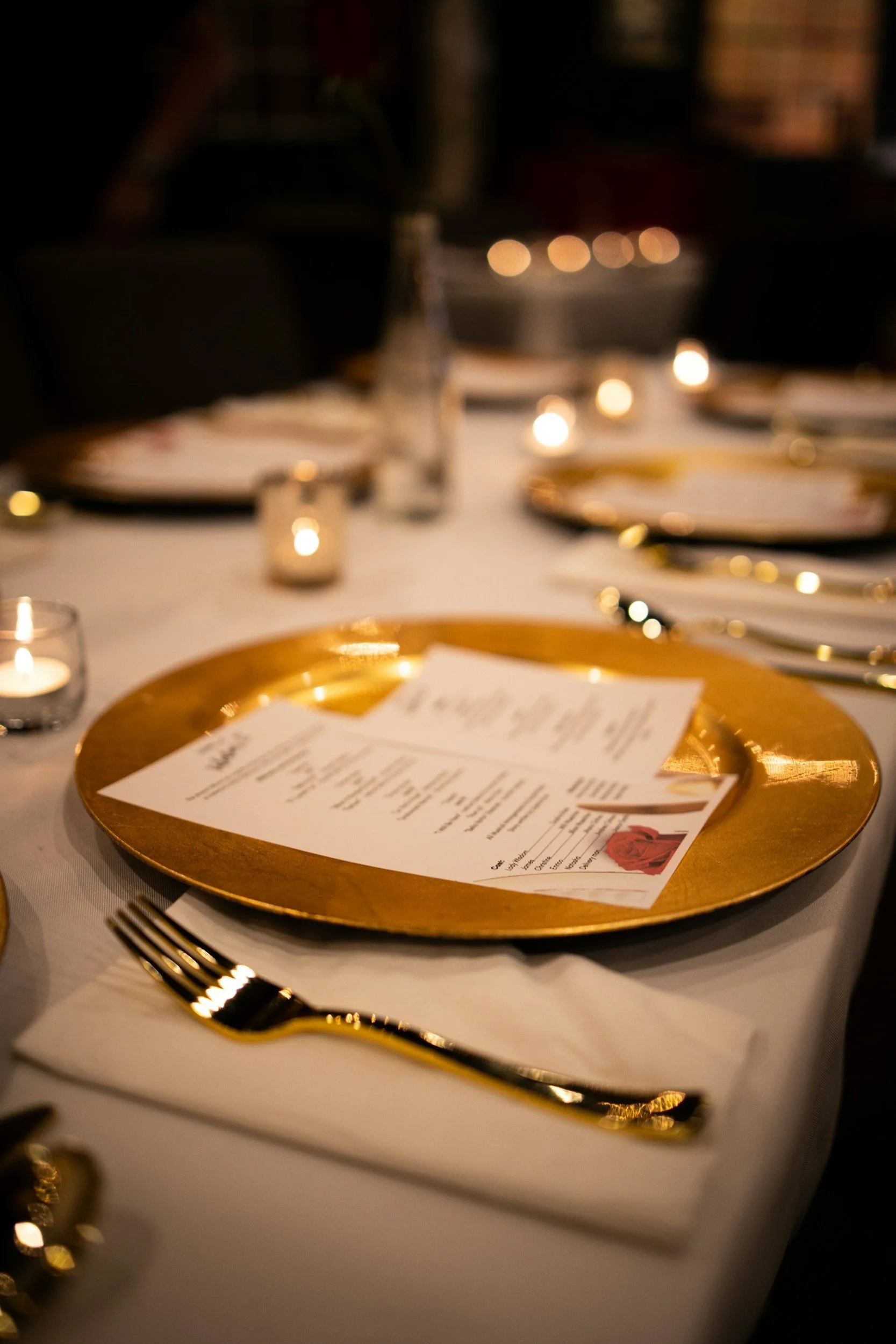 Elegant dinner table setting with a gold charger plate, printed menu, white napkin, and gold cutlery. Soft candle lighting and blurred background create a cozy ambiance.