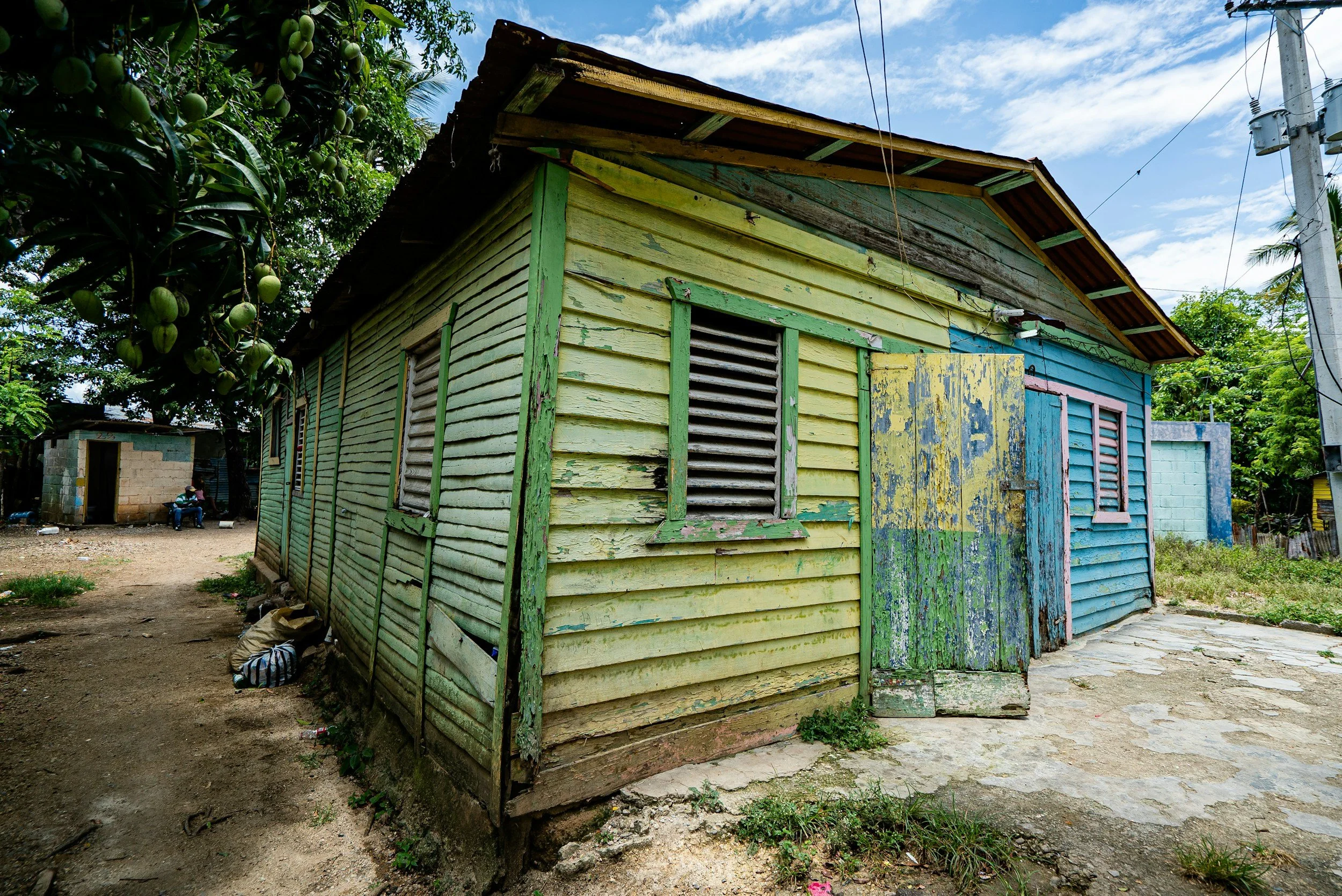 An old, weathered wooden house with peeling yellow and blue paint, surrounded by a dirt yard and green trees, under a partly cloudy sky.