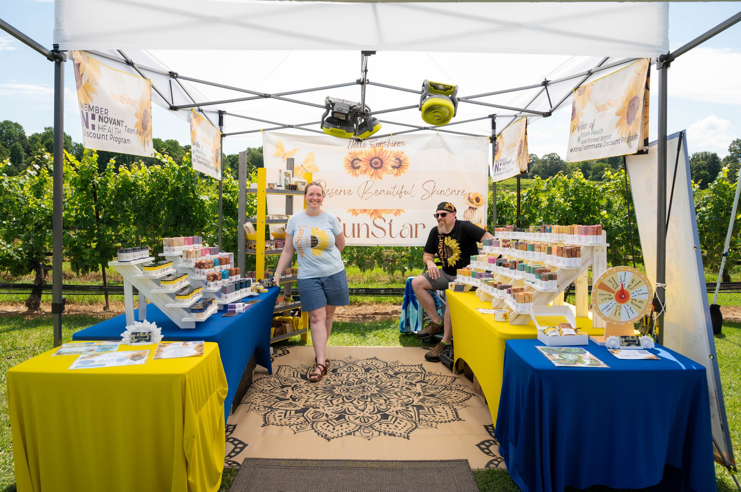 A vendor booth at an outdoor fair selling skincare products with the owner and her husband smiling; a woman standing and a man sitting, in front of a vineyard, under a canopy with sunflower banners.