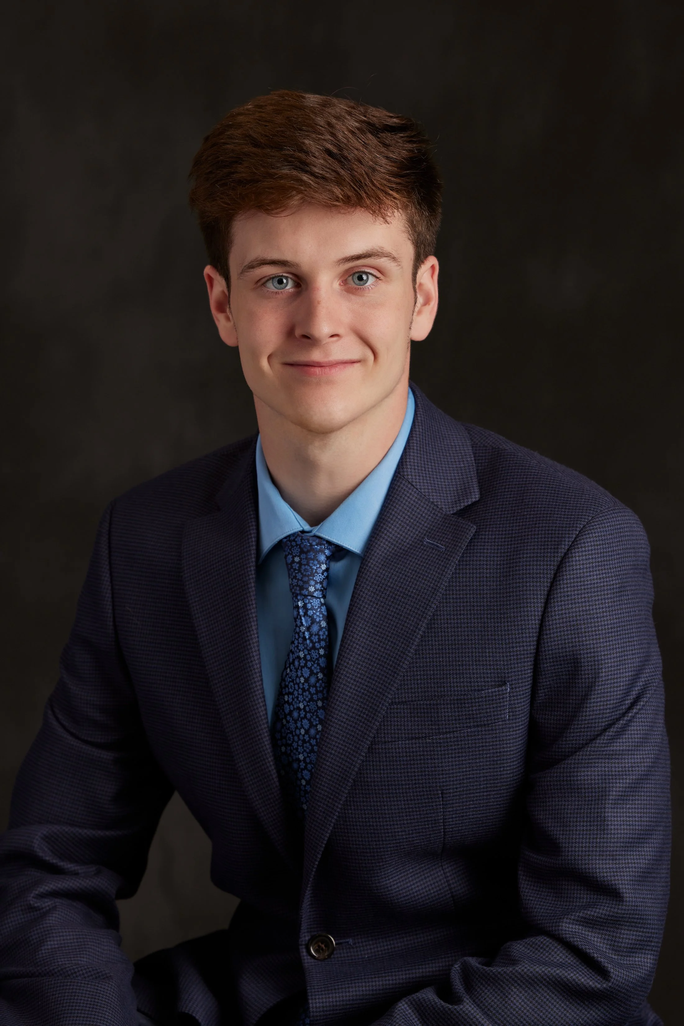 Professional portrait of a young man with light skin, short brown hair, wearing a navy blue checkered suit, light blue dress shirt, and patterned blue tie against a dark background.