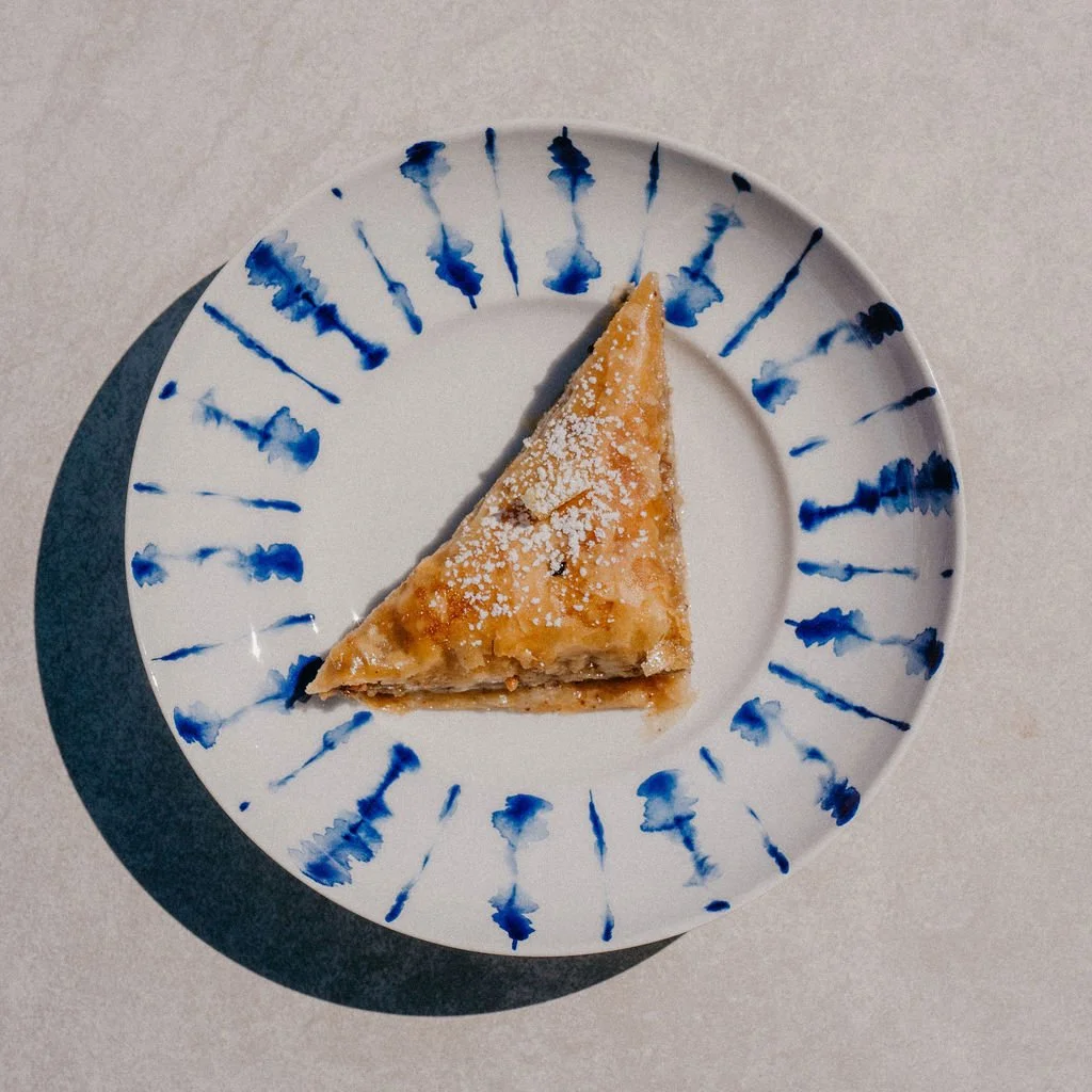 A slice of apple pie dusted with powdered sugar on a white plate with blue decorative streaks, placed on a light gray surface.