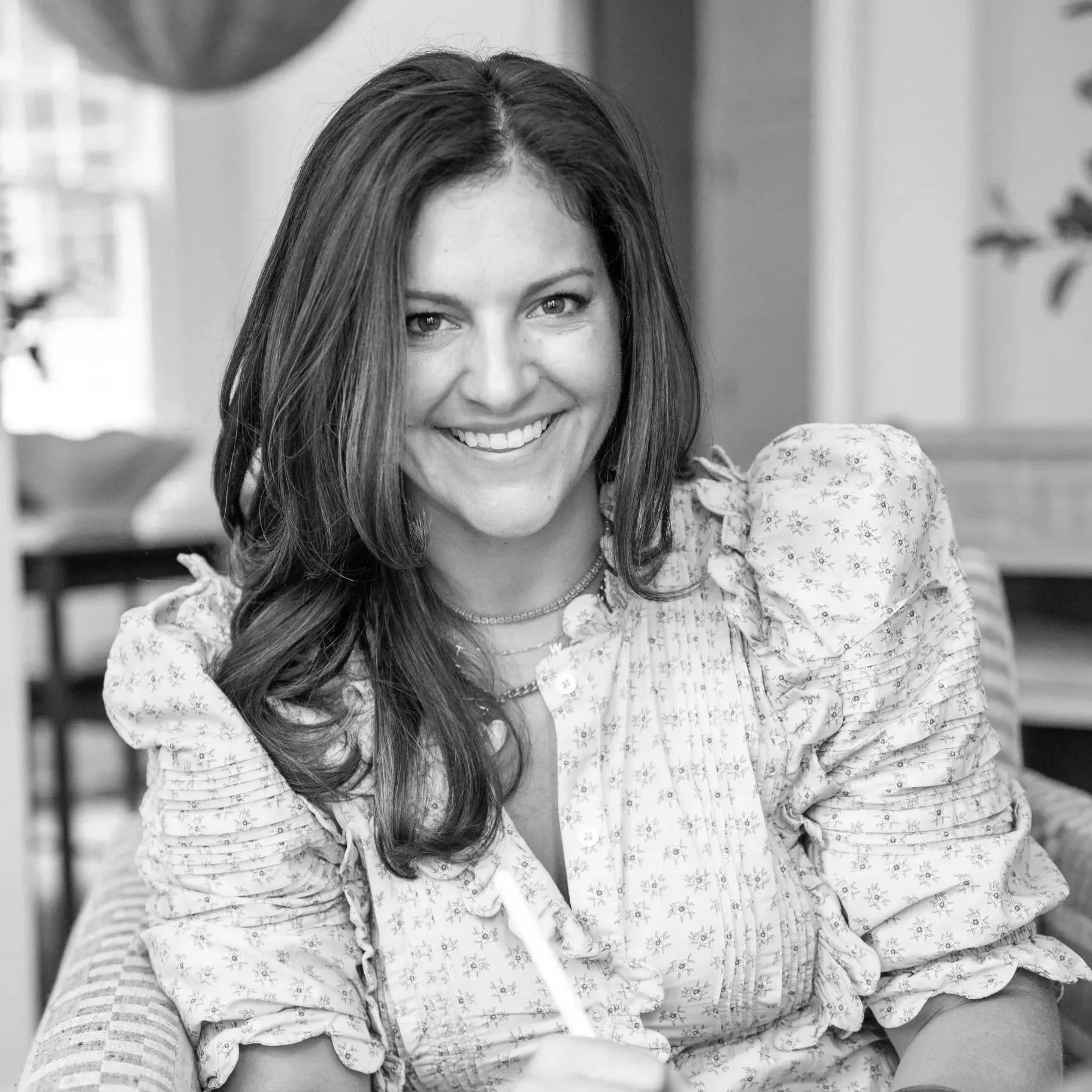 A smiling woman with long, wavy hair, wearing a floral patterned blouse with ruffled sleeves, sitting indoors with a blurred background.