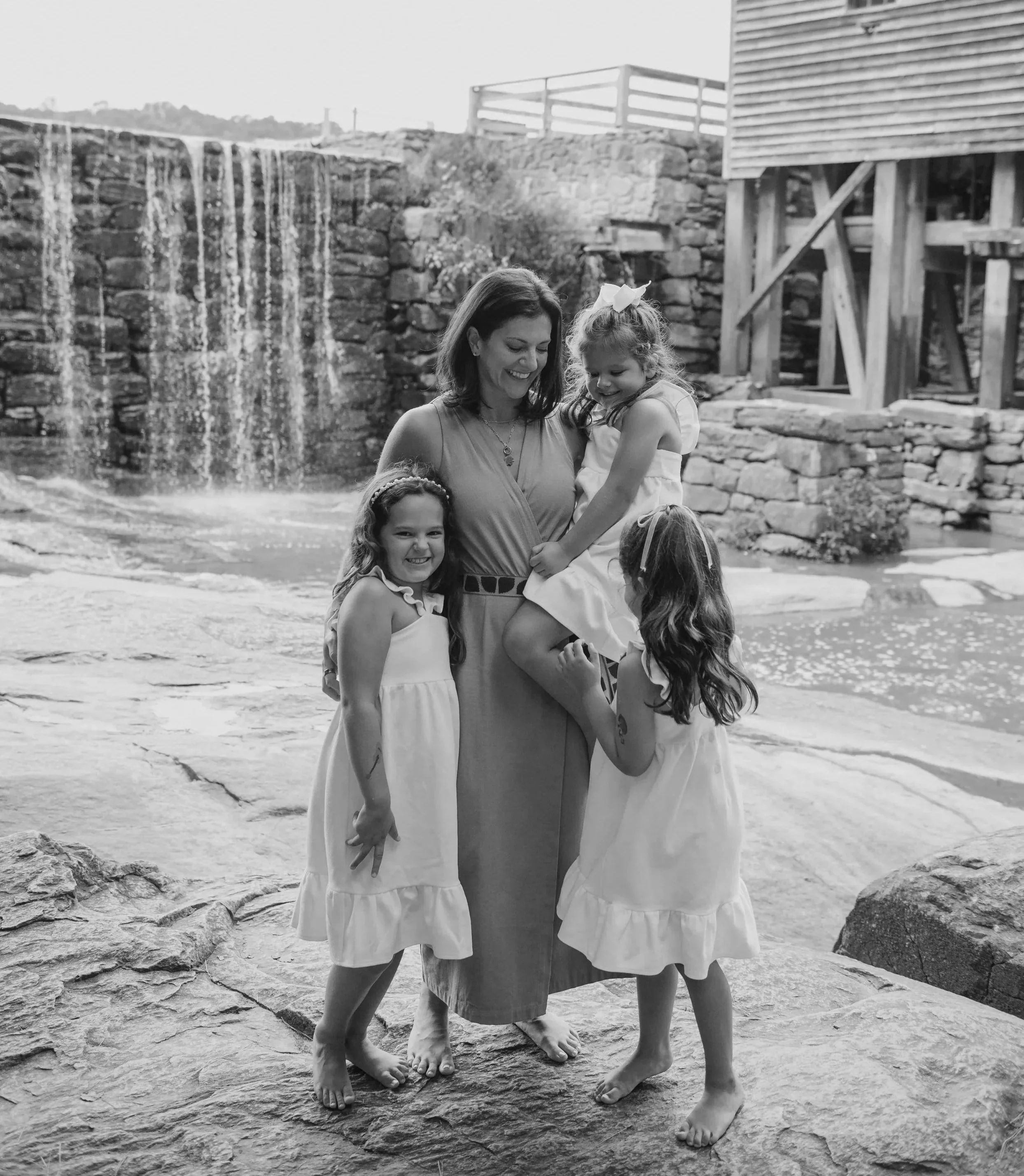 A woman and four young girls in matching white dresses standing on rocks near a waterfall and river.