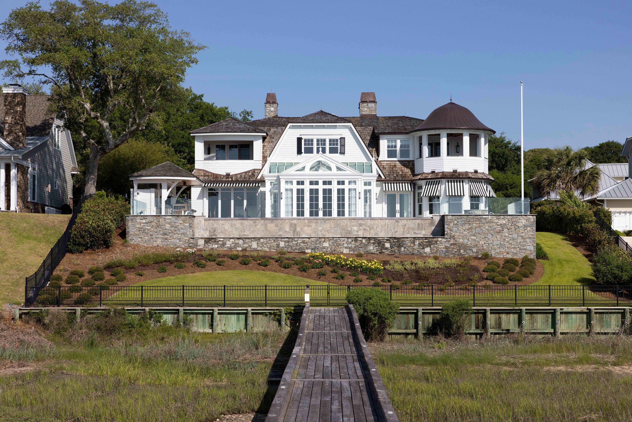 Large white Hamptons-style house on Bald Eagle Lane, Wilmington, NC, with a wooden dock leading to it, surrounded by a grassy lawn and landscaping, under a clear blue sky.