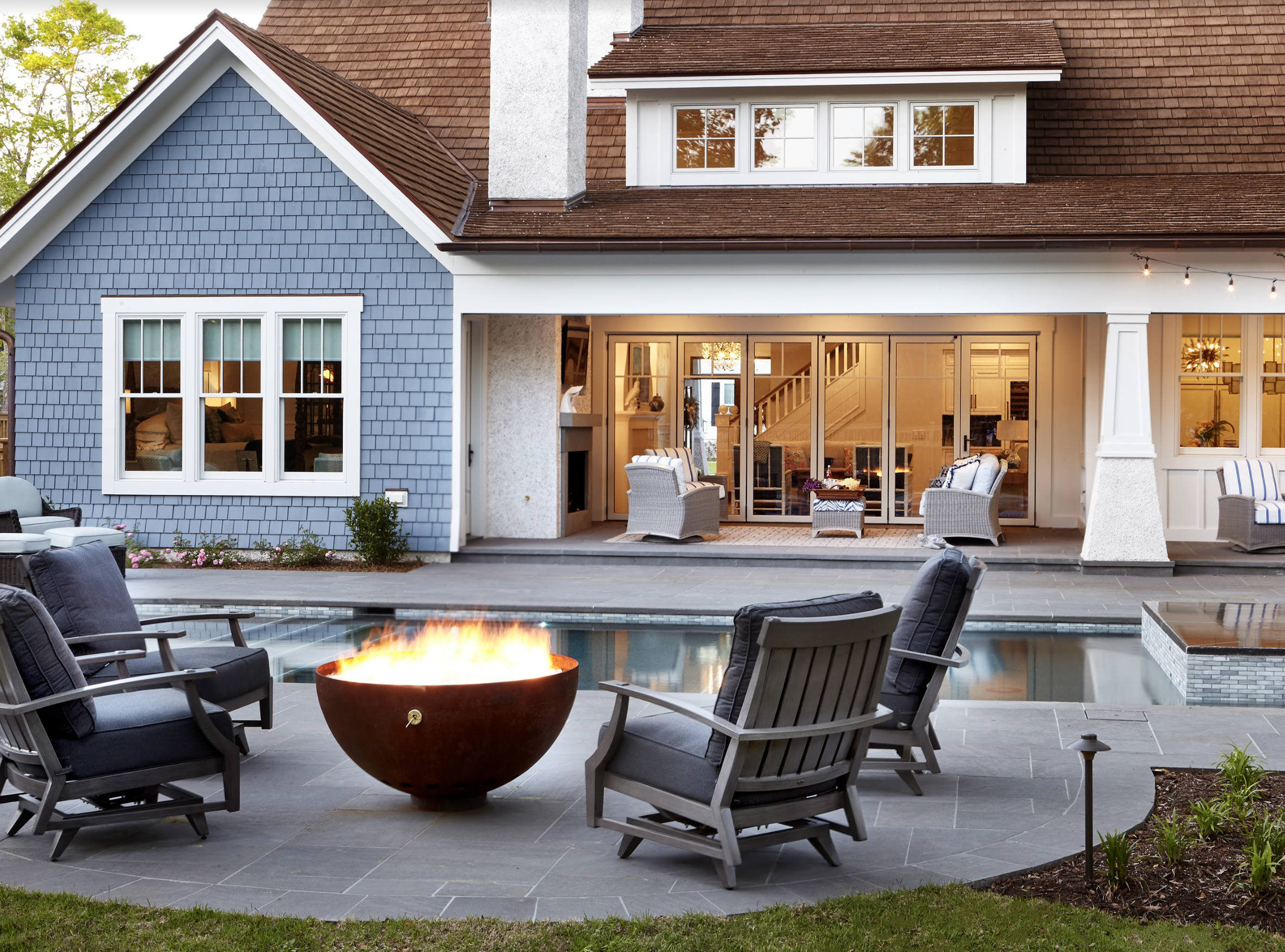 Backyard of a Raleigh, NC, house with a swimming pool, outdoor fire pit, patio seating, and a house with large windows, a chimney, and a covered porch area.