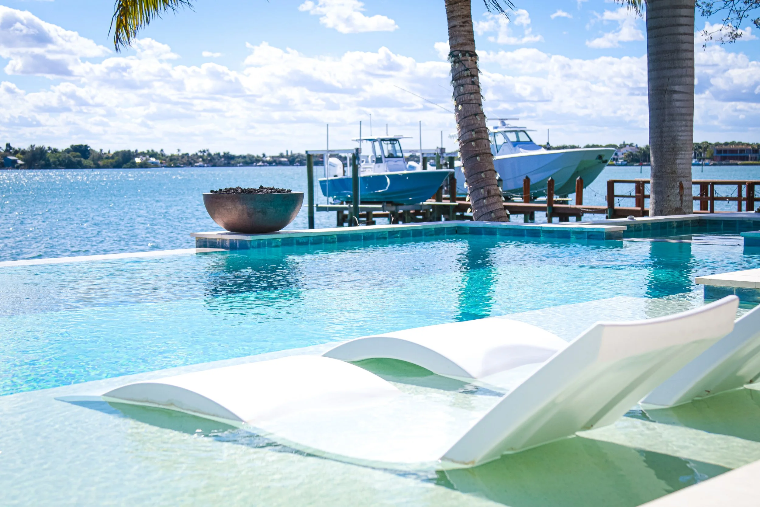 Aerial view of a modern Florida house with white roof, surrounded by palm trees, overlooking a green water body. There is a small swimming pool and a dock extending into the water.