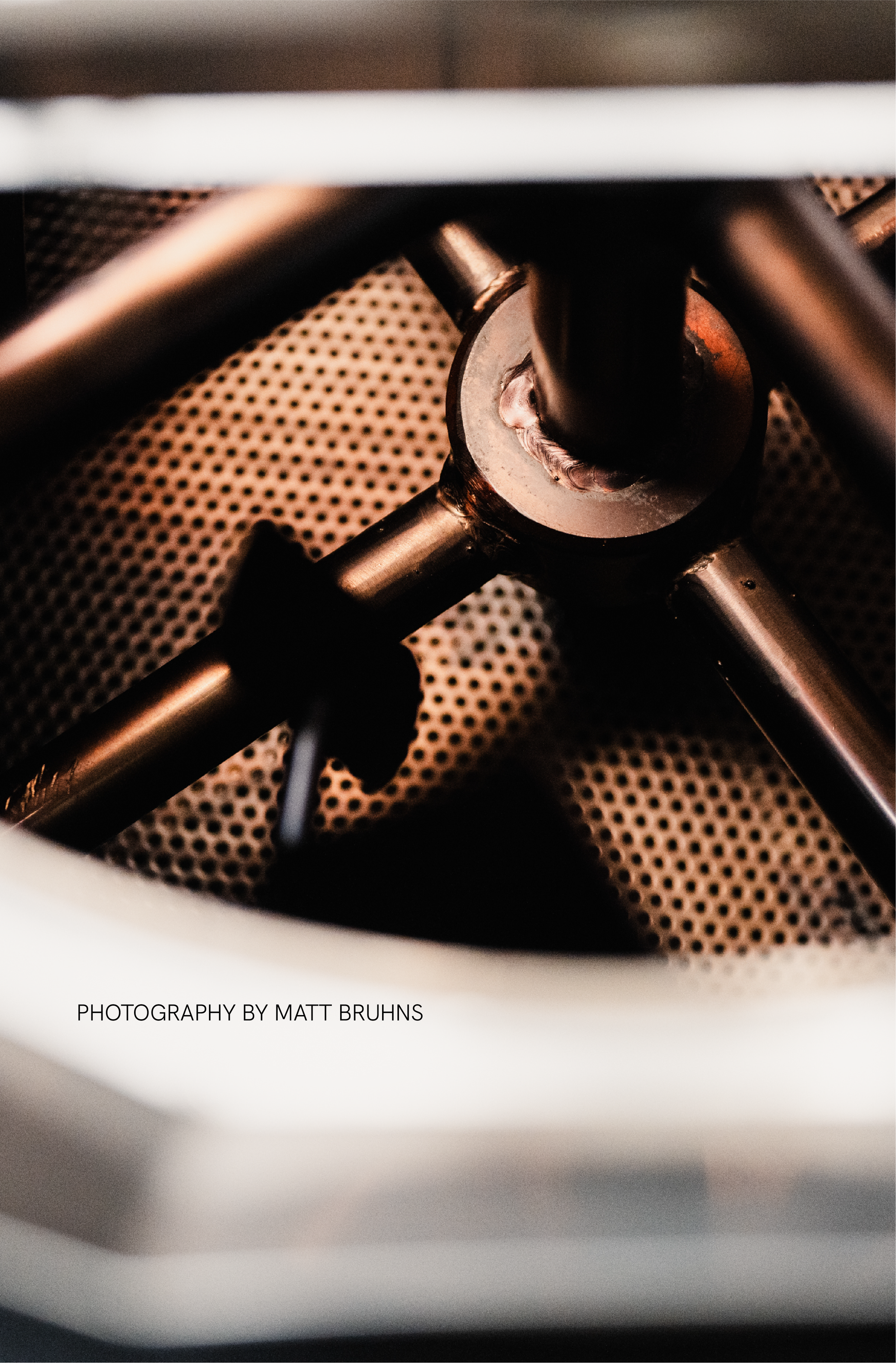 Close-up of a coffee roaster interior with metal arms and perforated surface, lit with warm tones, photographed by Matt Bruhns.