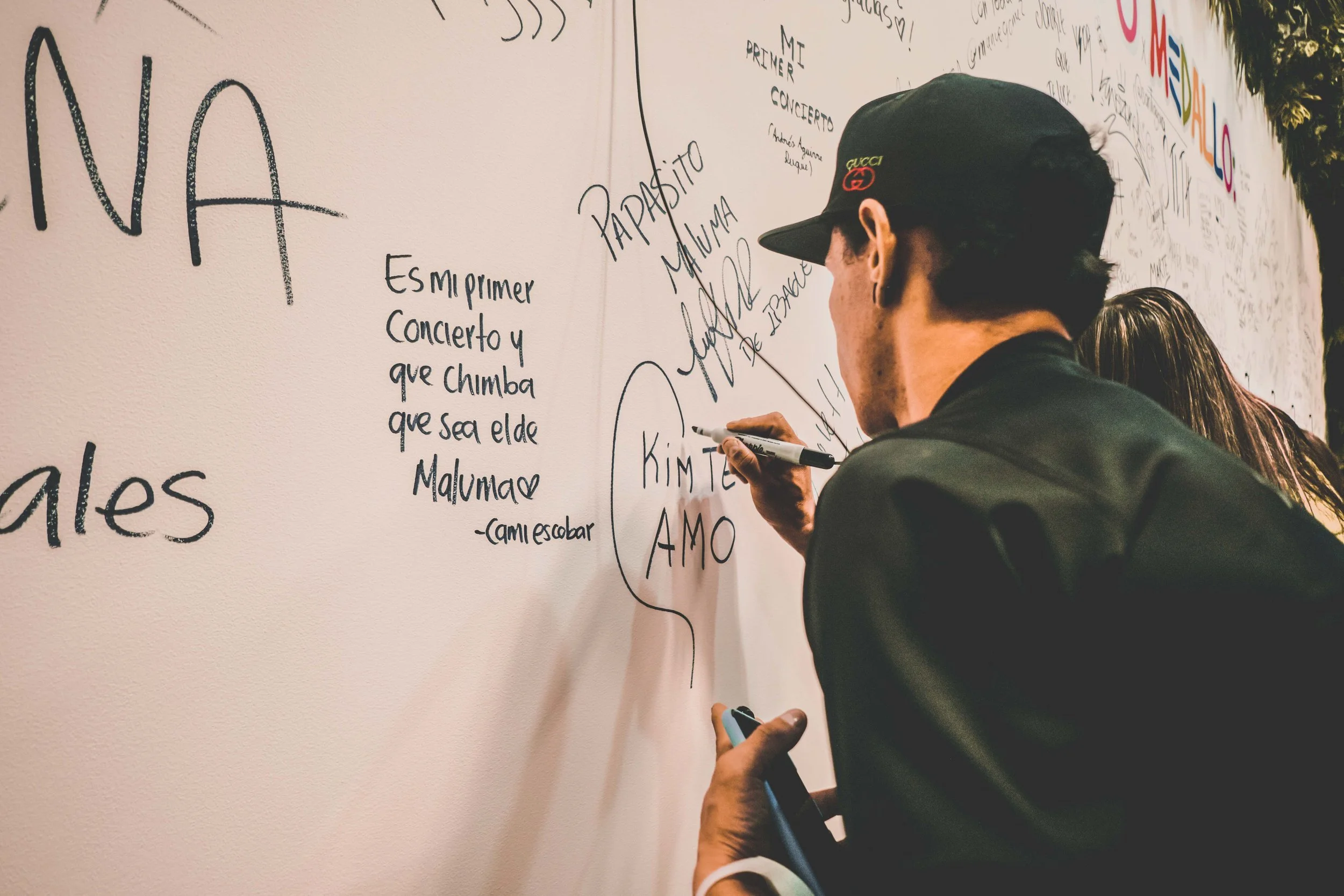 A man wearing a black cap and jacket writes on a white wall filled with various handwritten messages with a black marker.