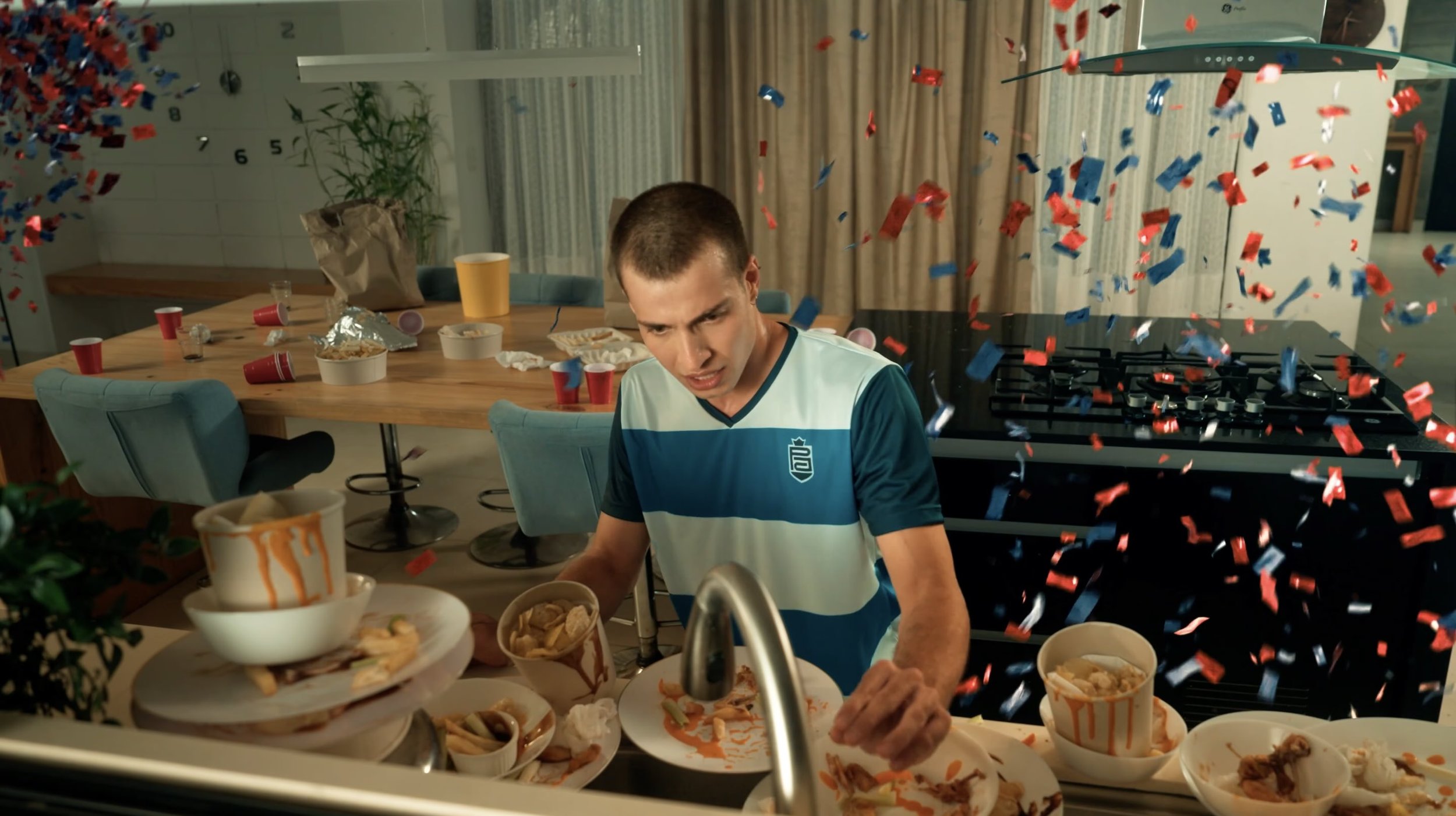 A young man with short brown hair in a blue and white striped sports shirt is cleaning up after a party, with red and blue confetti falling around him. The kitchen counter is cluttered with empty plates, bowls, and snack remnants, and the background shows a table with more party debris and a kitchen stovetop.