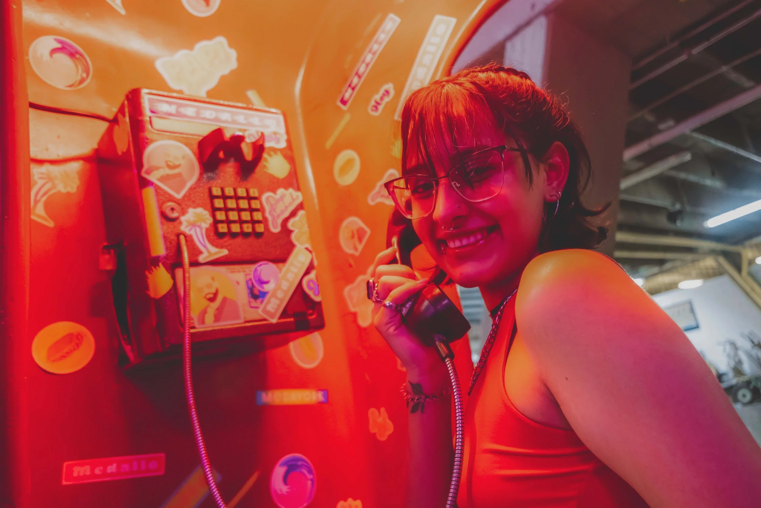 A woman with short hair, glasses, and a nose piercing smiling while talking on a payphone. She is wearing an orange tank top and standing next to a colorful wall with stickers and a payphone booth.