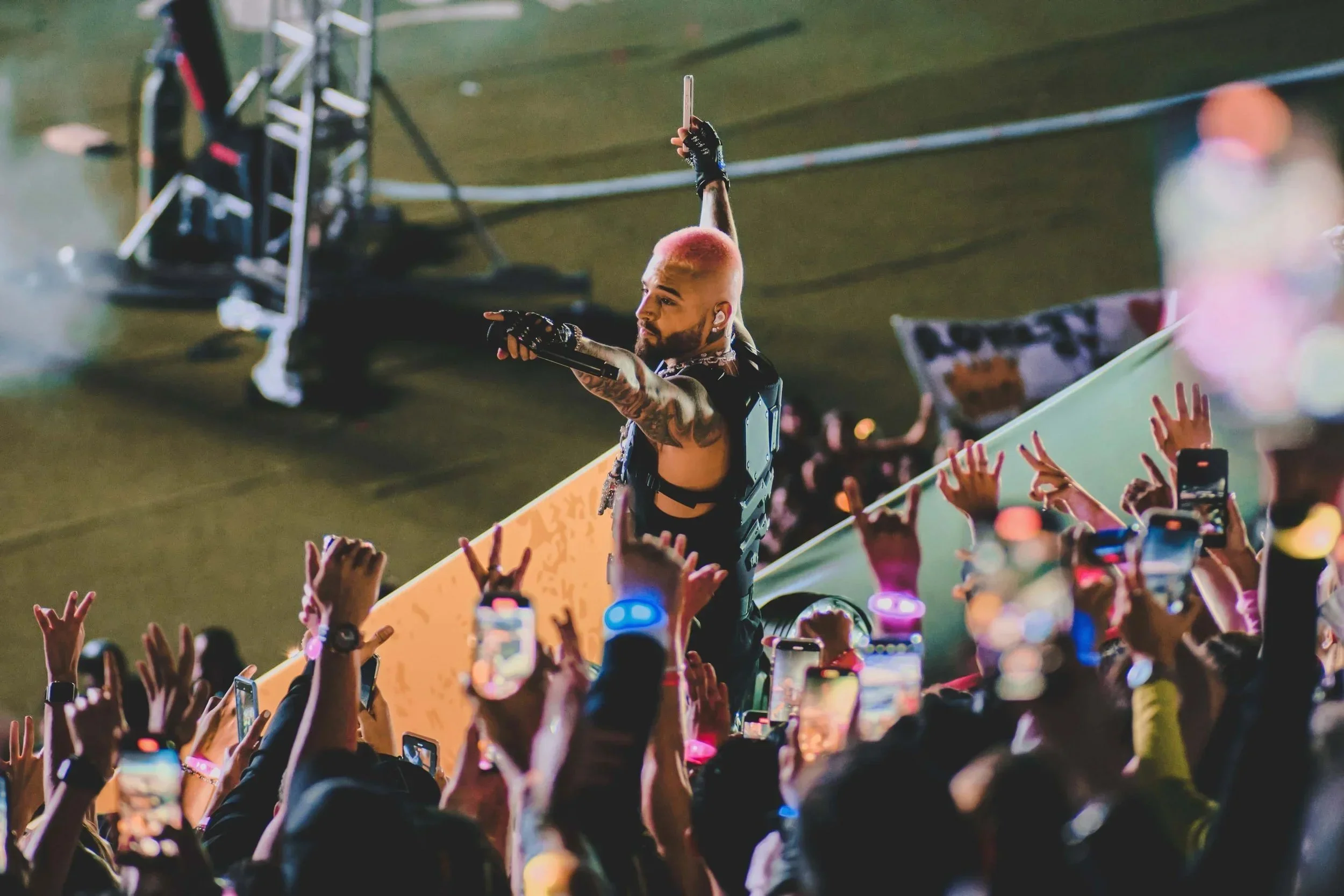 A performer with pink hair and tattoos on his arms is on stage, holding a phone high in the air. He is wearing black clothing with harnesses and gloves. The crowd in front of him is raising their hands, many holding phones to record the performance, and some are making hand gestures. The scene is lively with colorful lights and a blurred background.