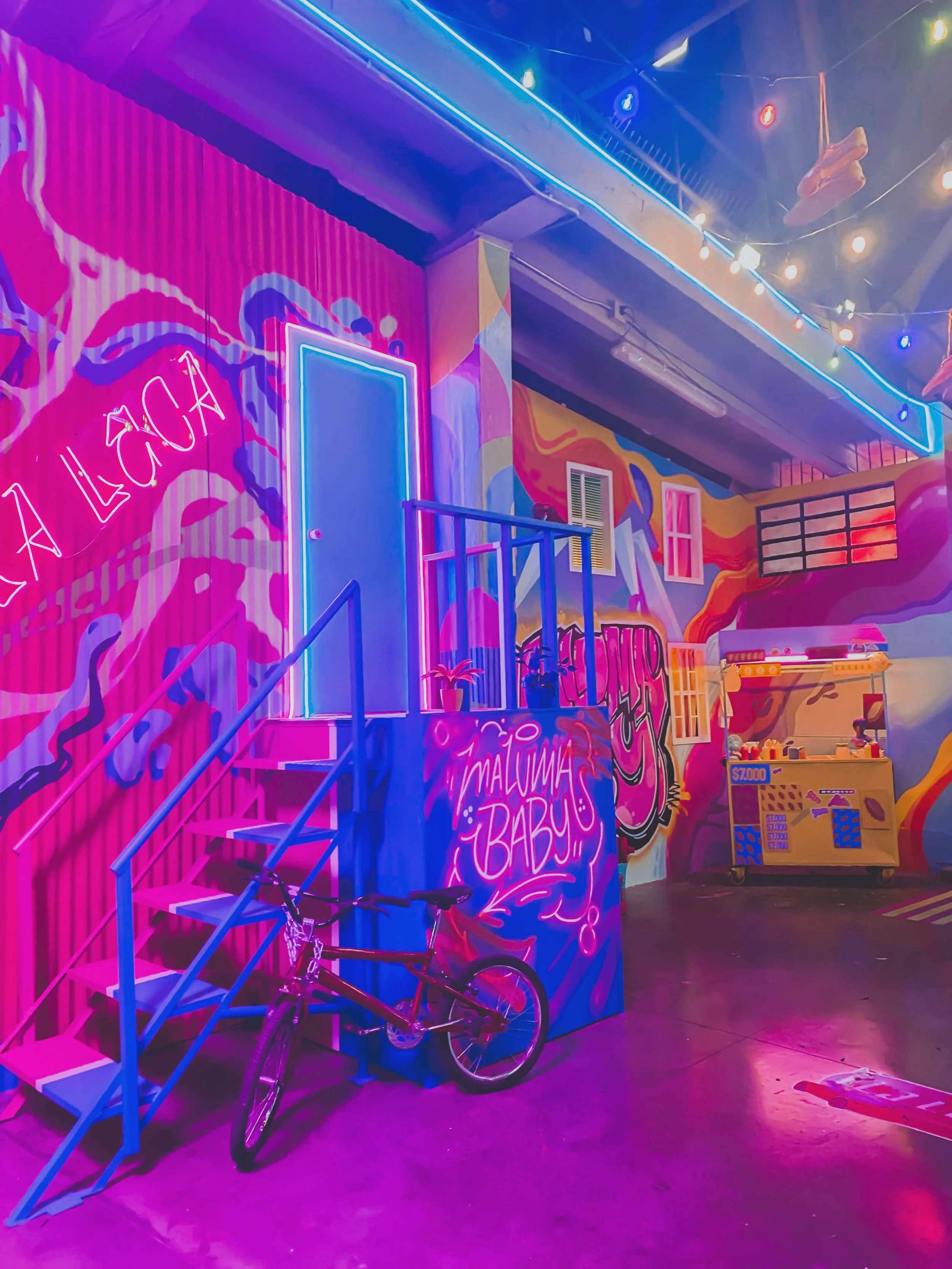 Colorful indoor space with vibrant neon lights, graffiti-style murals, and a small staircase leading to a closed blue door. There's a bicycle parked beside the stairs and an arcade game in the background.
