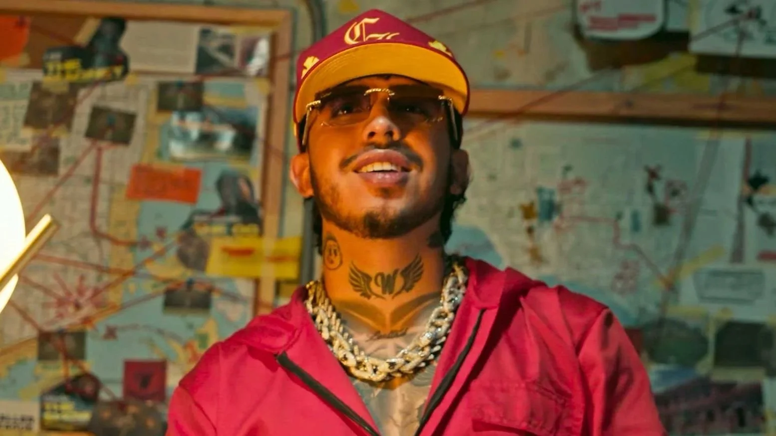 A man wearing sunglasses, a red cap, and a red jacket, with tattoos and jewelry, standing in front of a wall covered with maps and photographs.