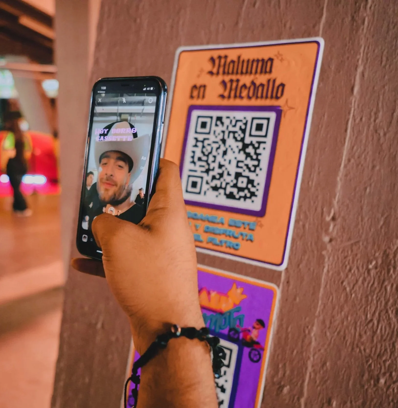 A person taking a selfie with a smartphone in front of a QR code sign on a wall. The person is smiling and wearing a black bracelet, and a woman with a yellow and green shirt is visible in the background.
