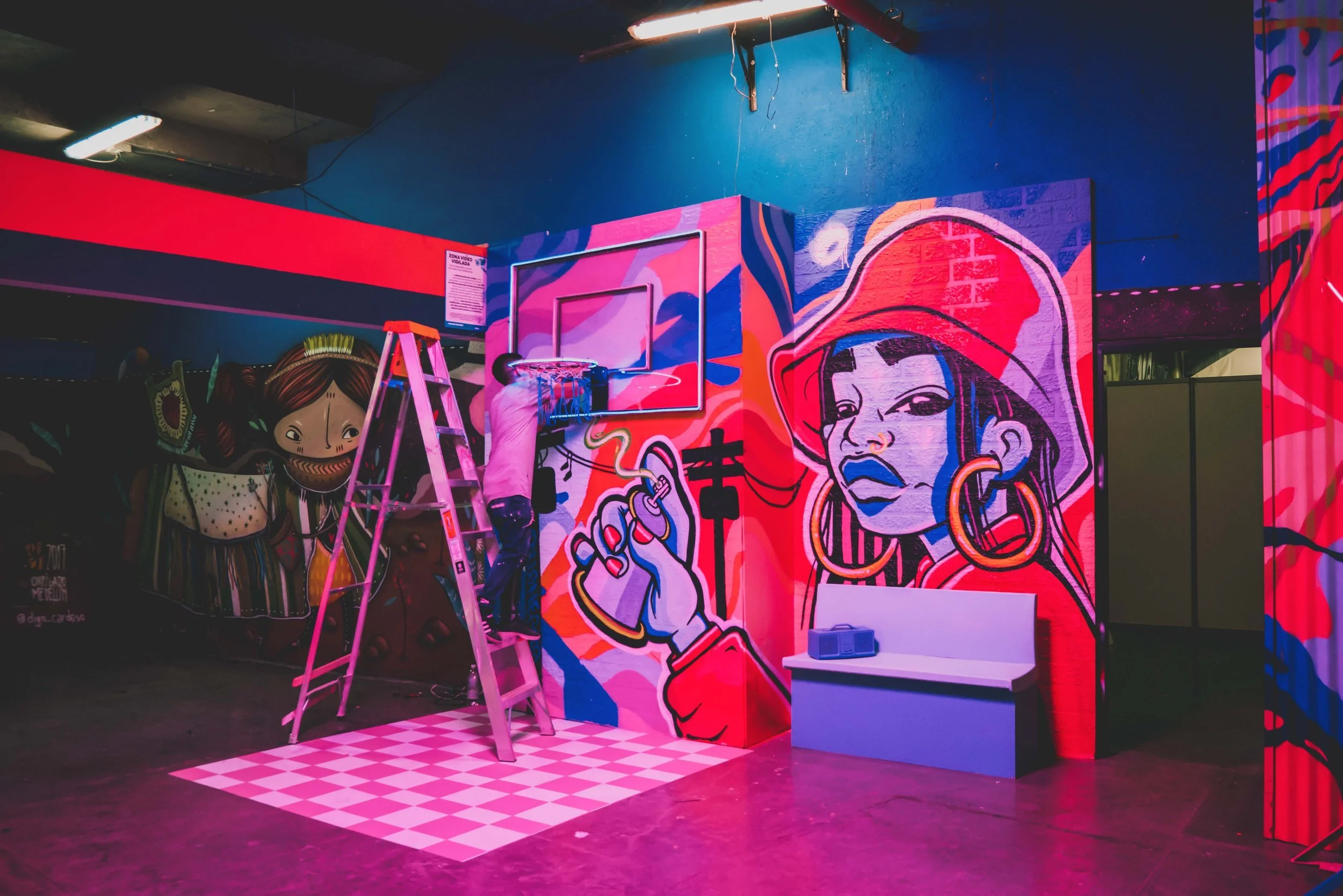 An indoor space with vibrant graffiti art on the walls, featuring a large feminine face holding a cigarette and a man installing a small basketball hoop onto a colorful painted wall. There is a ladder, a white bench, and a checkered pink and white floor section.