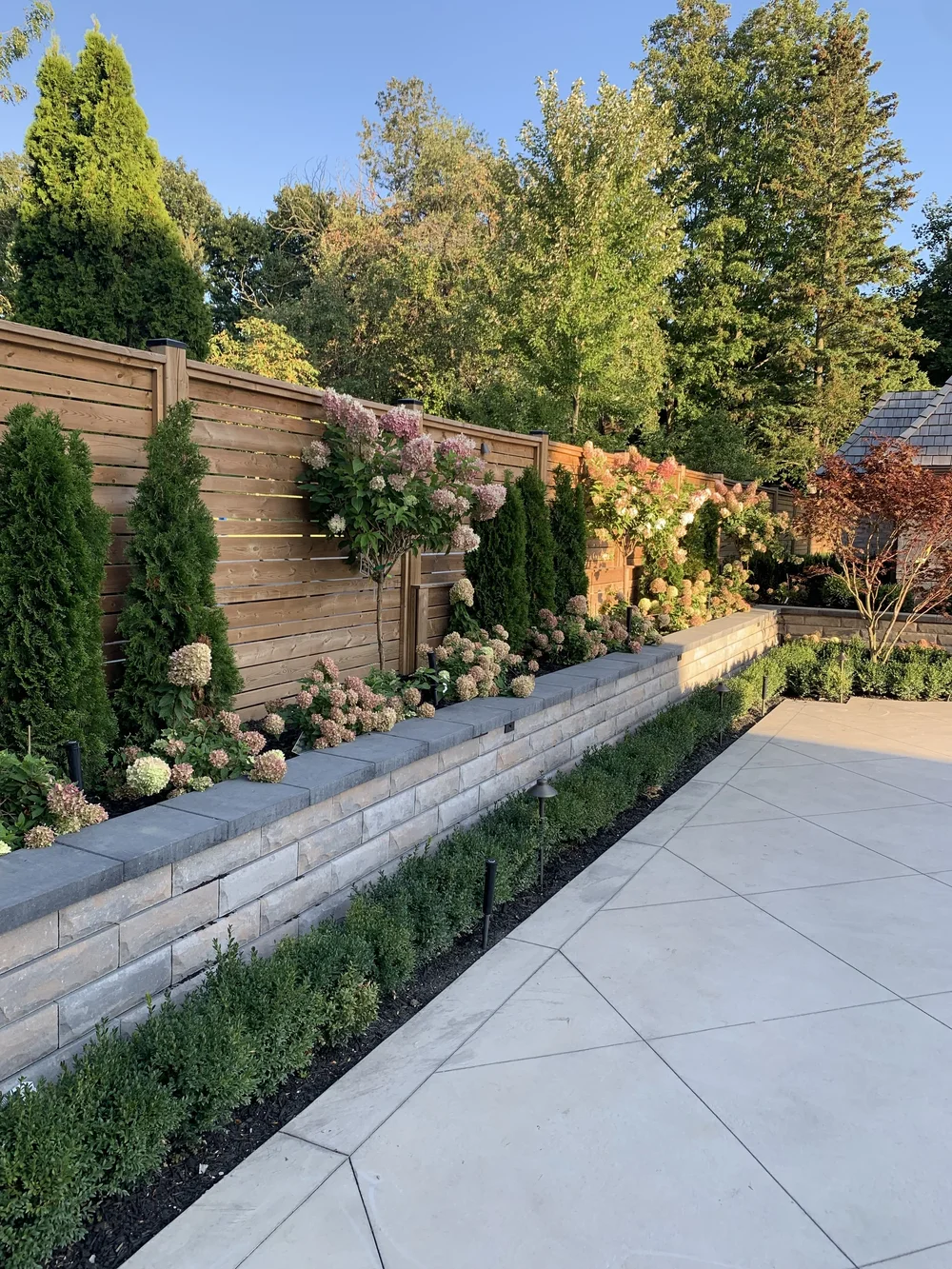 Stone retaining wall garden bed beside a large-format slab patio and cedar fence