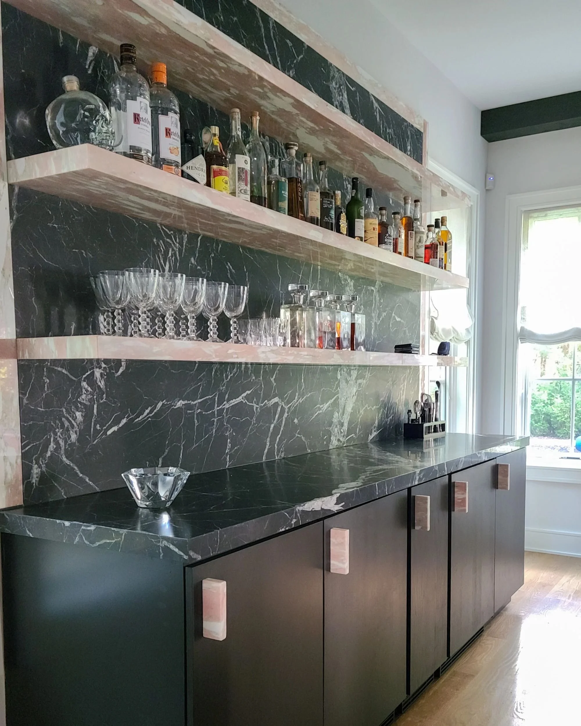 A bar with black marble countertop and matching marble backsplash, featuring open shelves with various liquor bottles and glassware, and below cabinet storage in black with pink marble handles, near a window with sunlight.