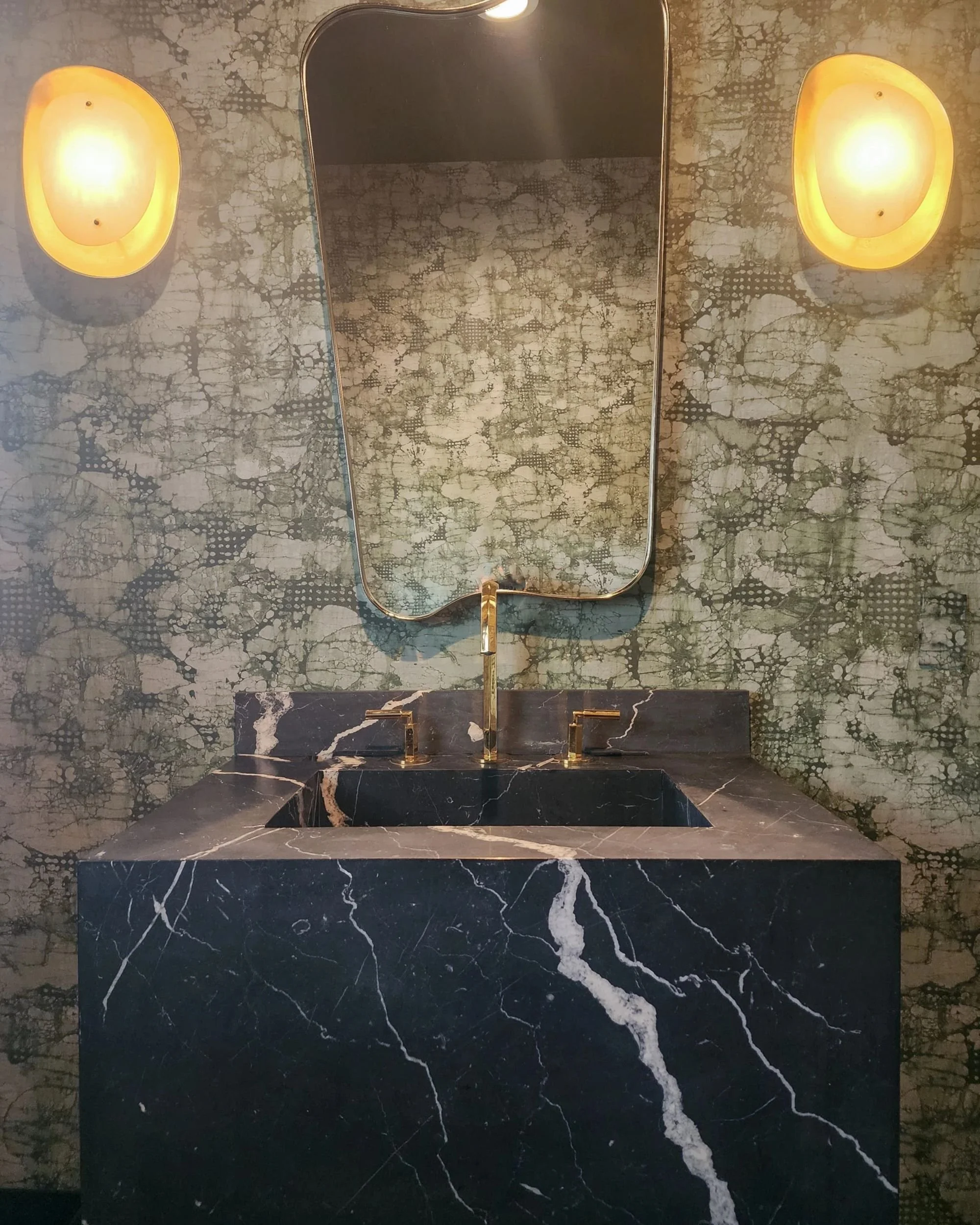 A bathroom vanity with a black marble sink and gold faucet, a large mirror above it, and two wall-mounted yellowish lights flanking the mirror.