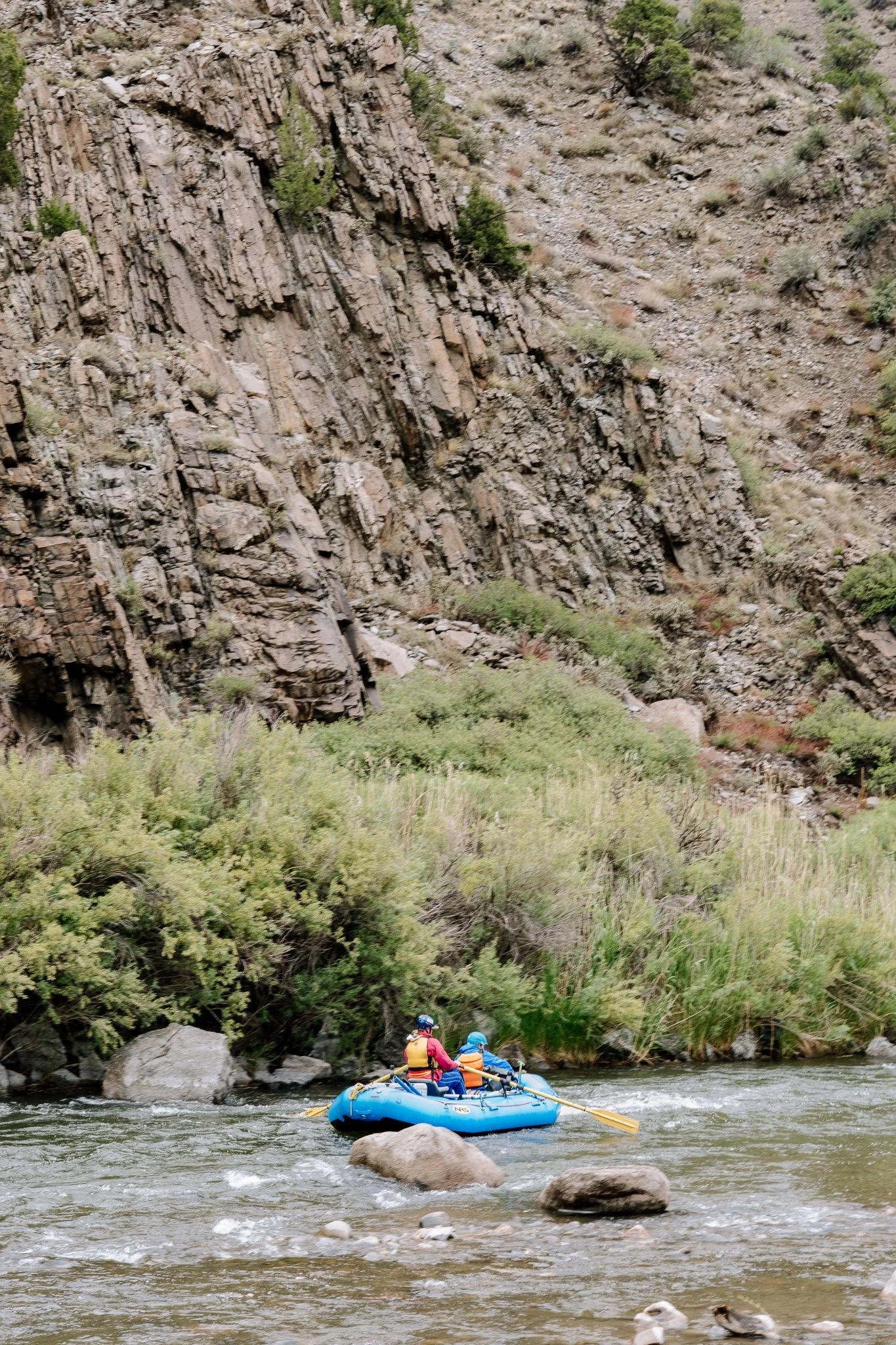 Two people in helmets and life jackets are paddling a blue inflatable raft on a river, with rocky terrain and green bushes along the riverbank.