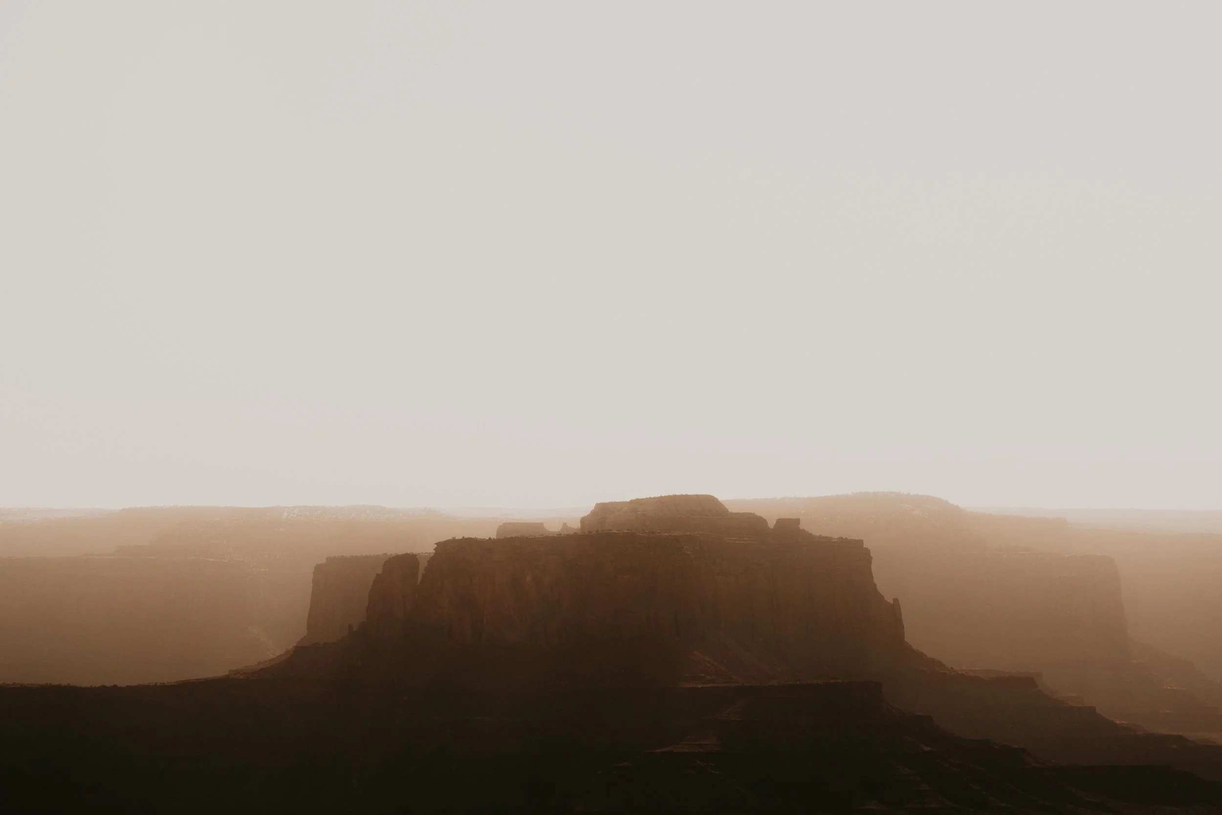 A landscape view of a canyon during sunset or sunrise, with mesas and cliffs silhouetted against a hazy sky.