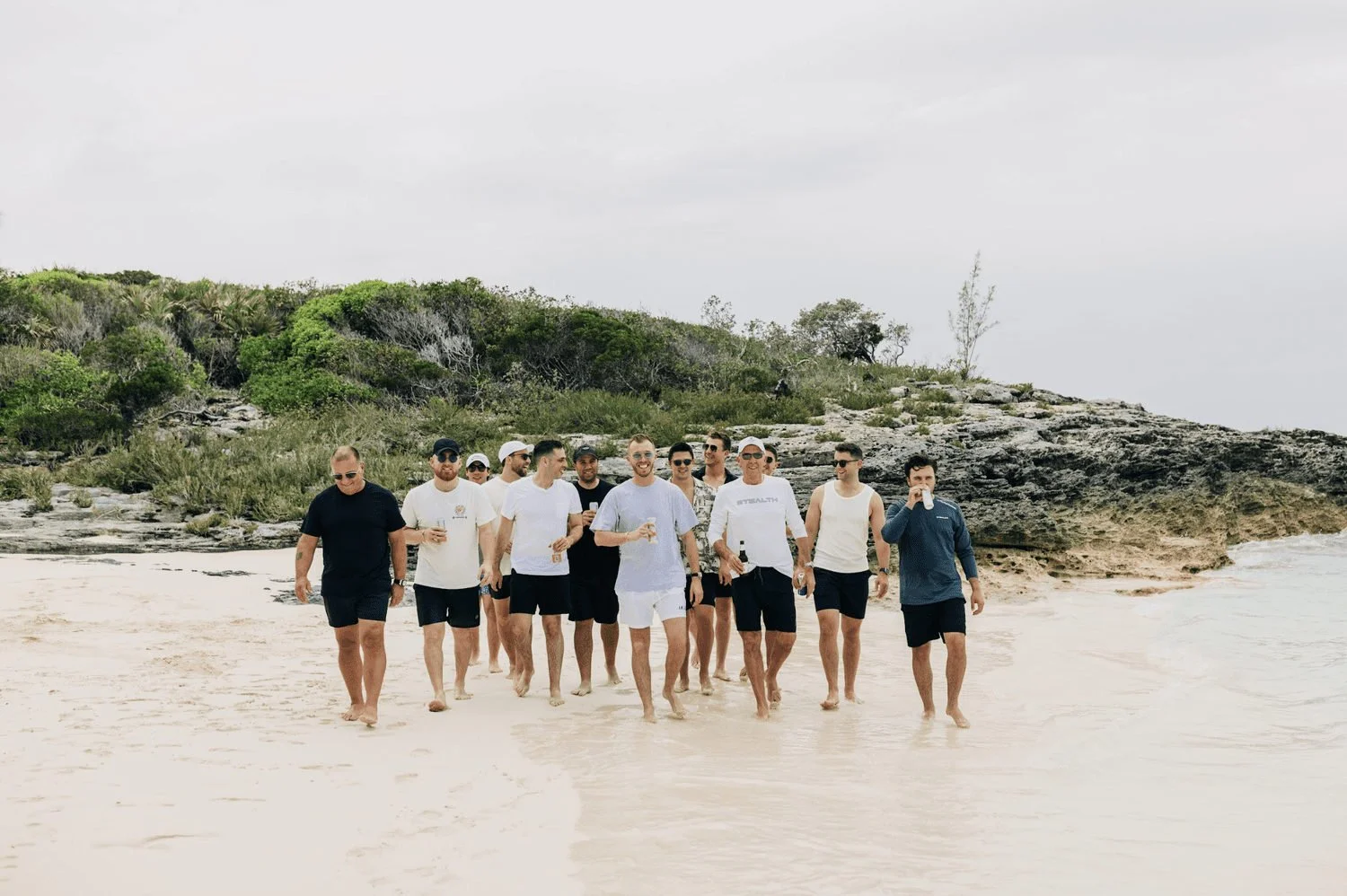 Bachelor party group walking together on a sandy beach beside a rocky shoreline.