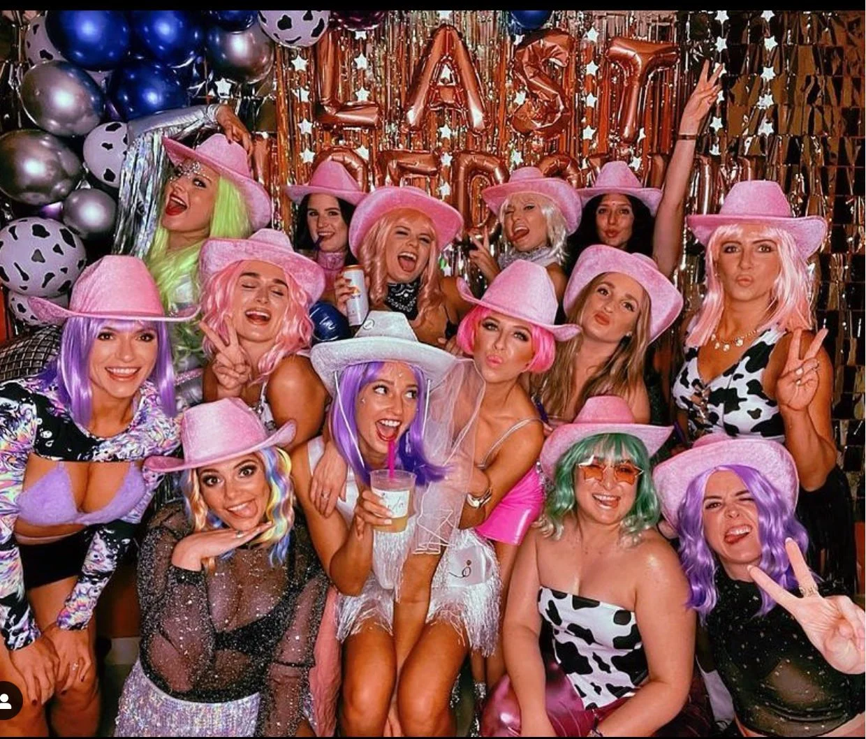 Bachelorette group in pink cowboy hats celebrating beneath balloons and letter decorations.