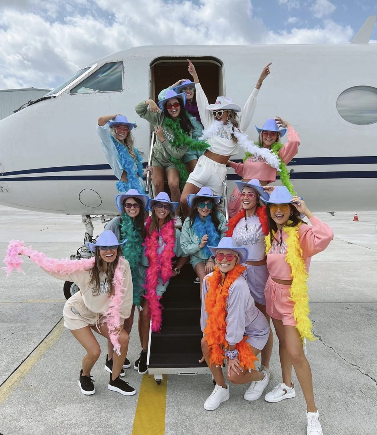 Friends in colorful outfits and feather boas posing beside a private jet before a bachelorette trip.