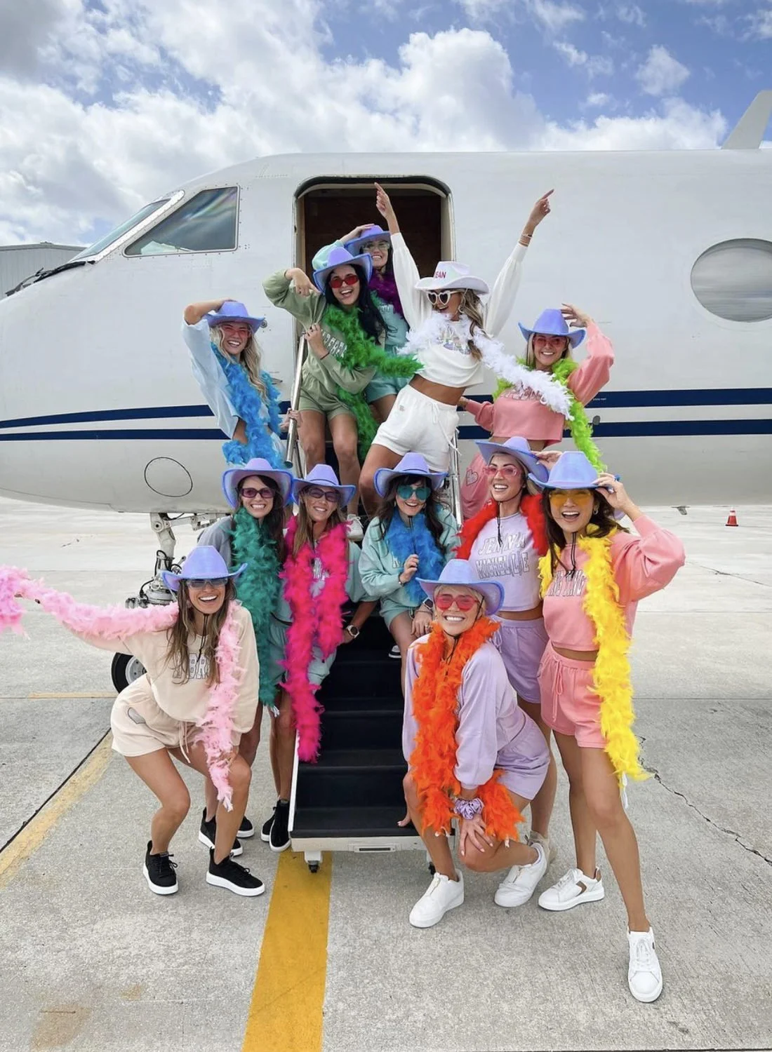 Friends in colorful outfits and feather boas posing beside a private jet before a bachelorette trip.