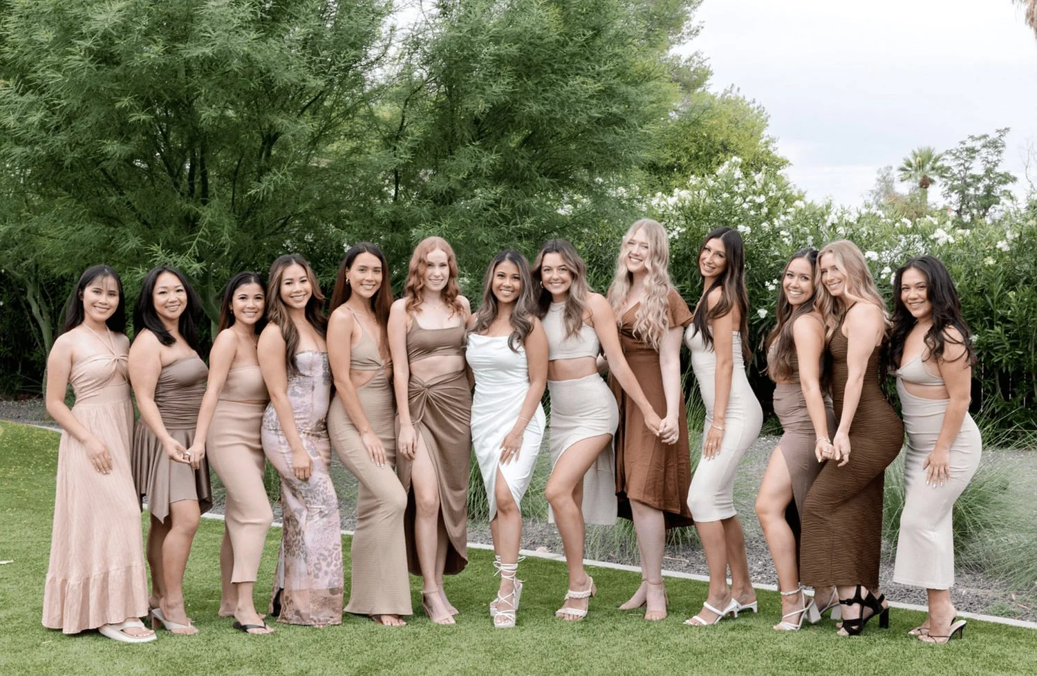 Bachelorette group in neutral-colored dresses posing together on a lawn.