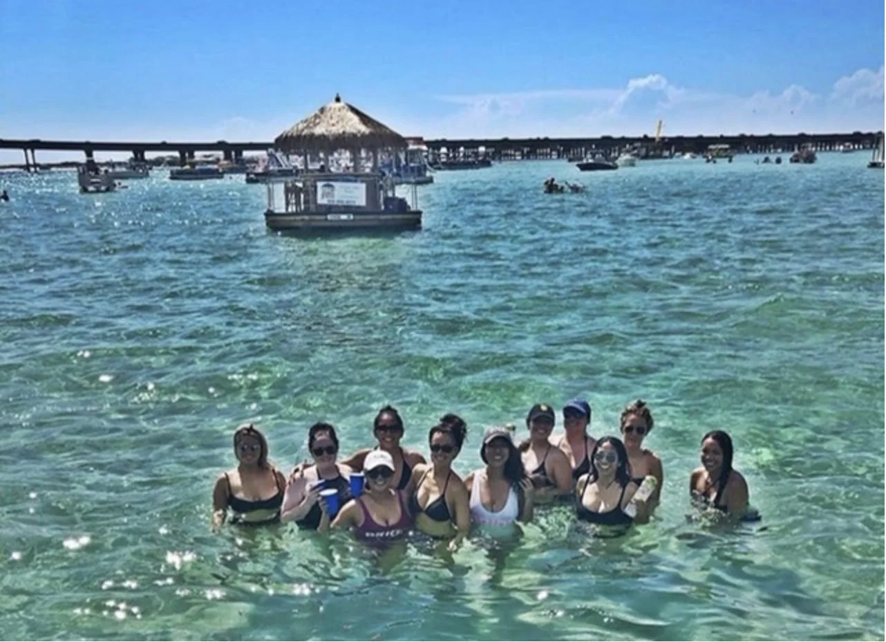 Bachelorette group standing together in shallow turquoise water near a floating tiki hut.