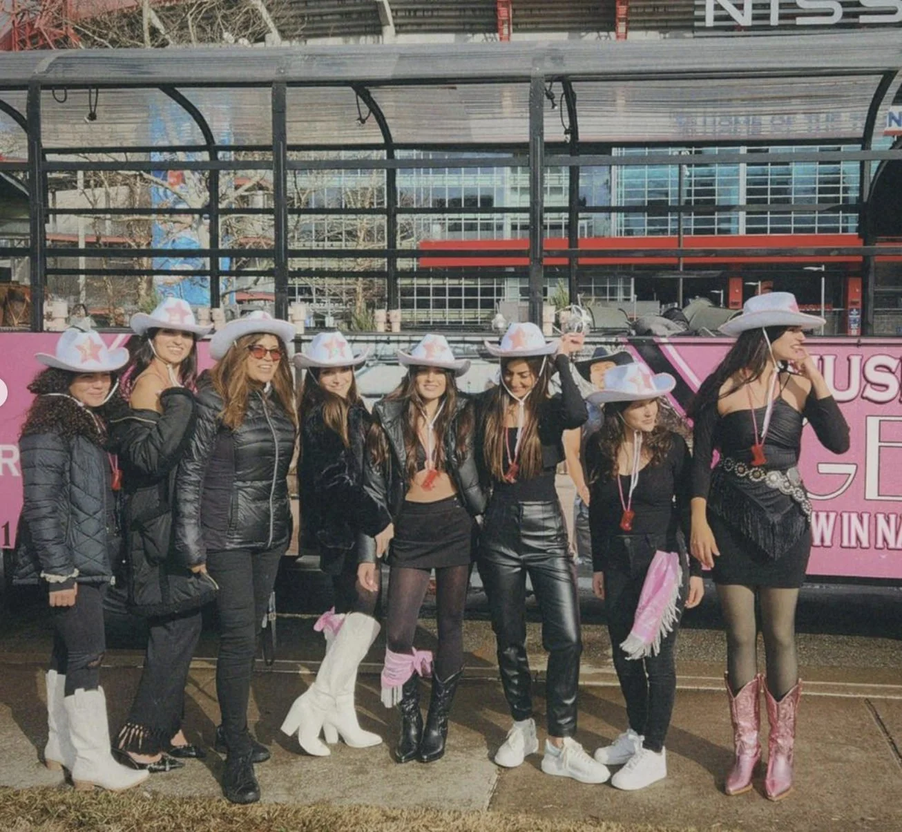 Bachelorette group in black outfits and matching cowboy hats posing beside a pink party bus in Nashville.