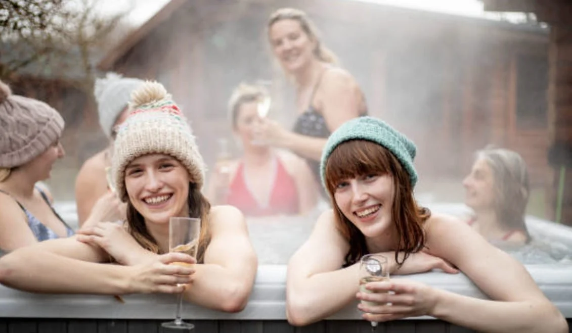 Friends wearing winter hats and holding drinks in an outdoor hot tub.