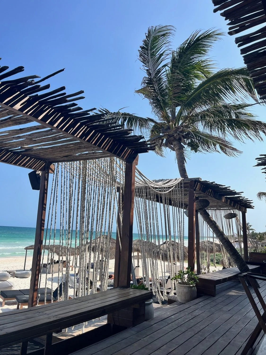Wooden beach deck with rope screens and palm trees overlooking turquoise water at Amansala in Tulum.