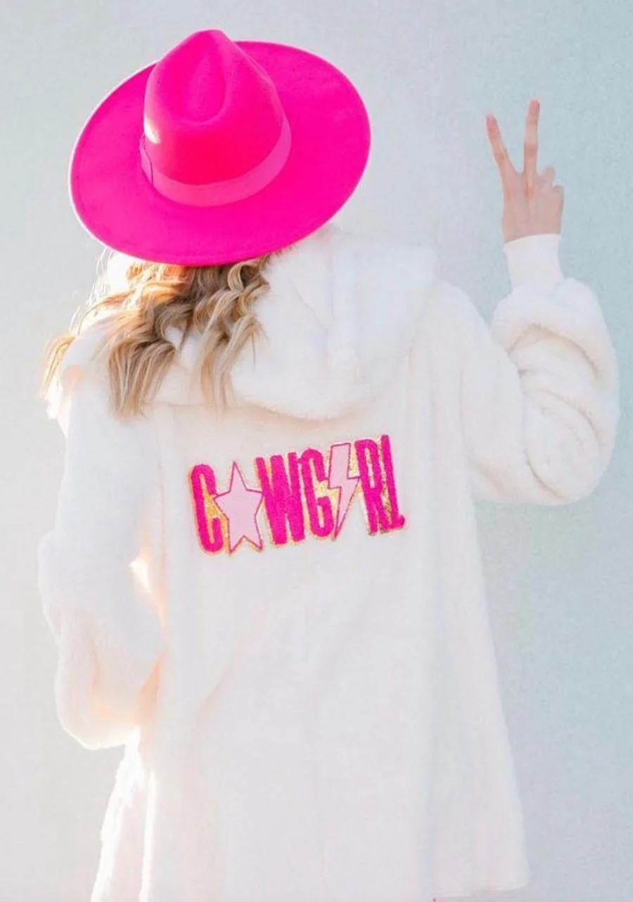 Woman in a bright pink cowboy hat wearing a white jacket with pink cowgirl lettering.