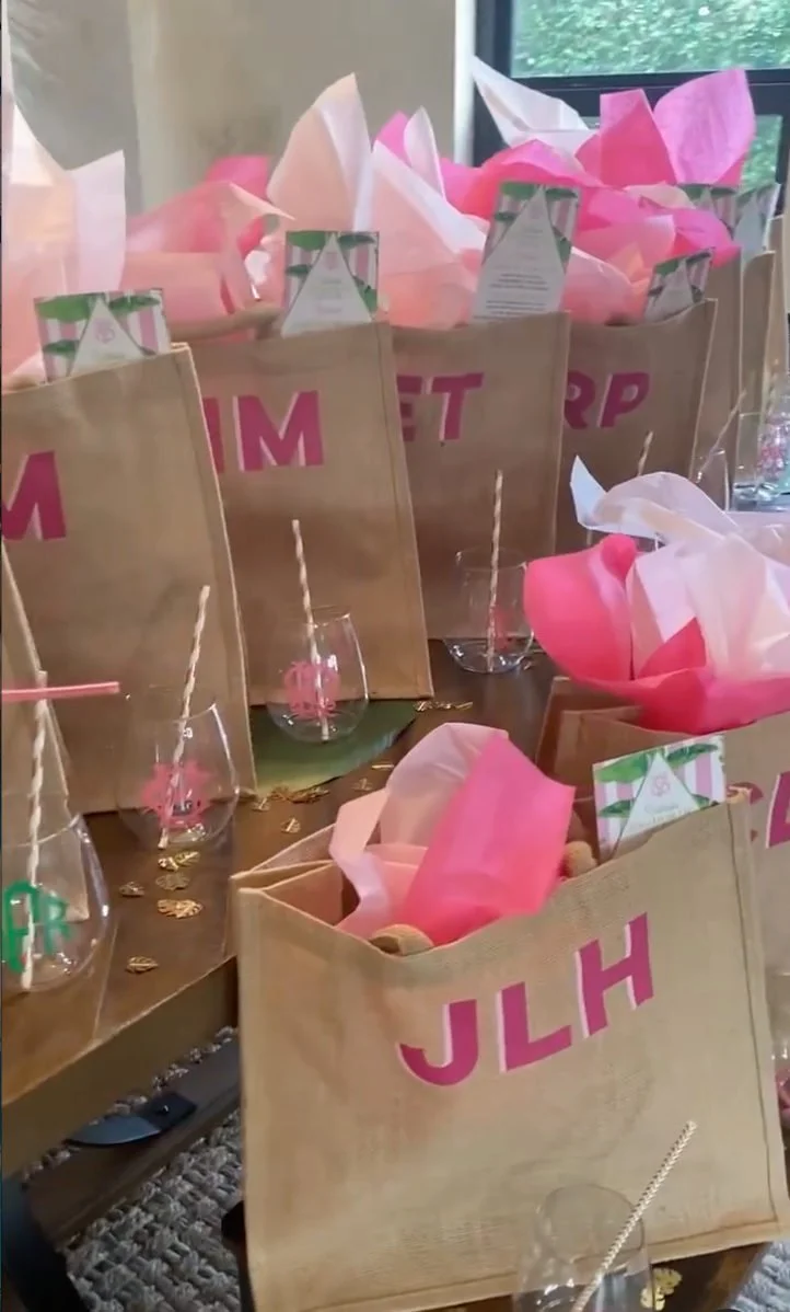 Personalized tote bags filled with pink tissue paper beside stemless party glasses.