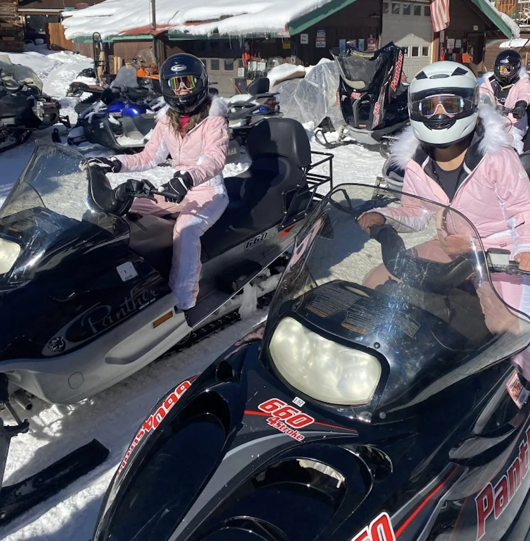 Friends wearing pink winter outfits and helmets seated on snowmobiles.