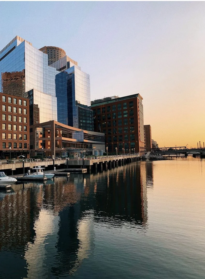 Boston waterfront buildings reflected in the harbor at sunset.