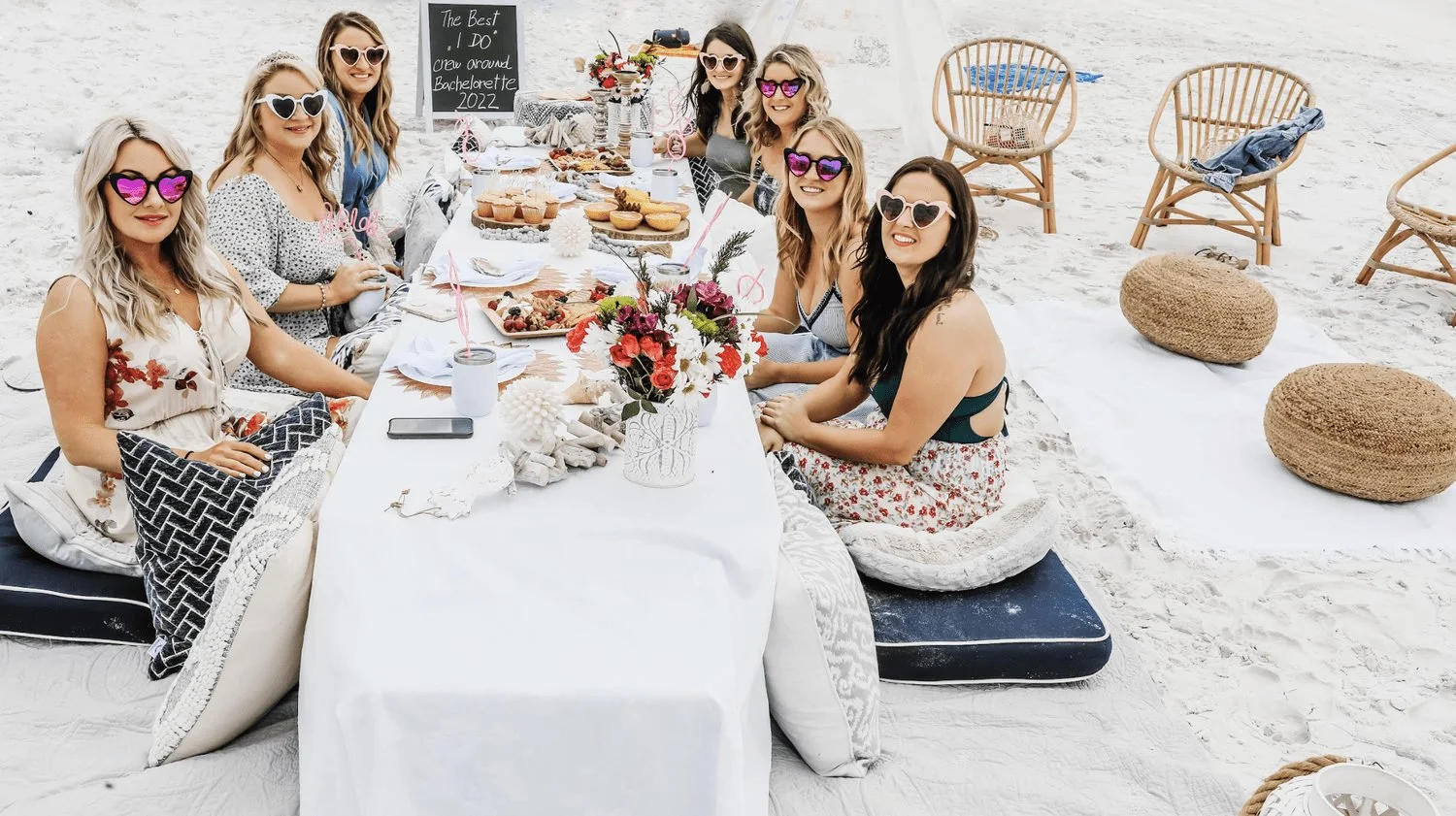 Bachelorette group seated around a decorated picnic table on a white-sand beach.