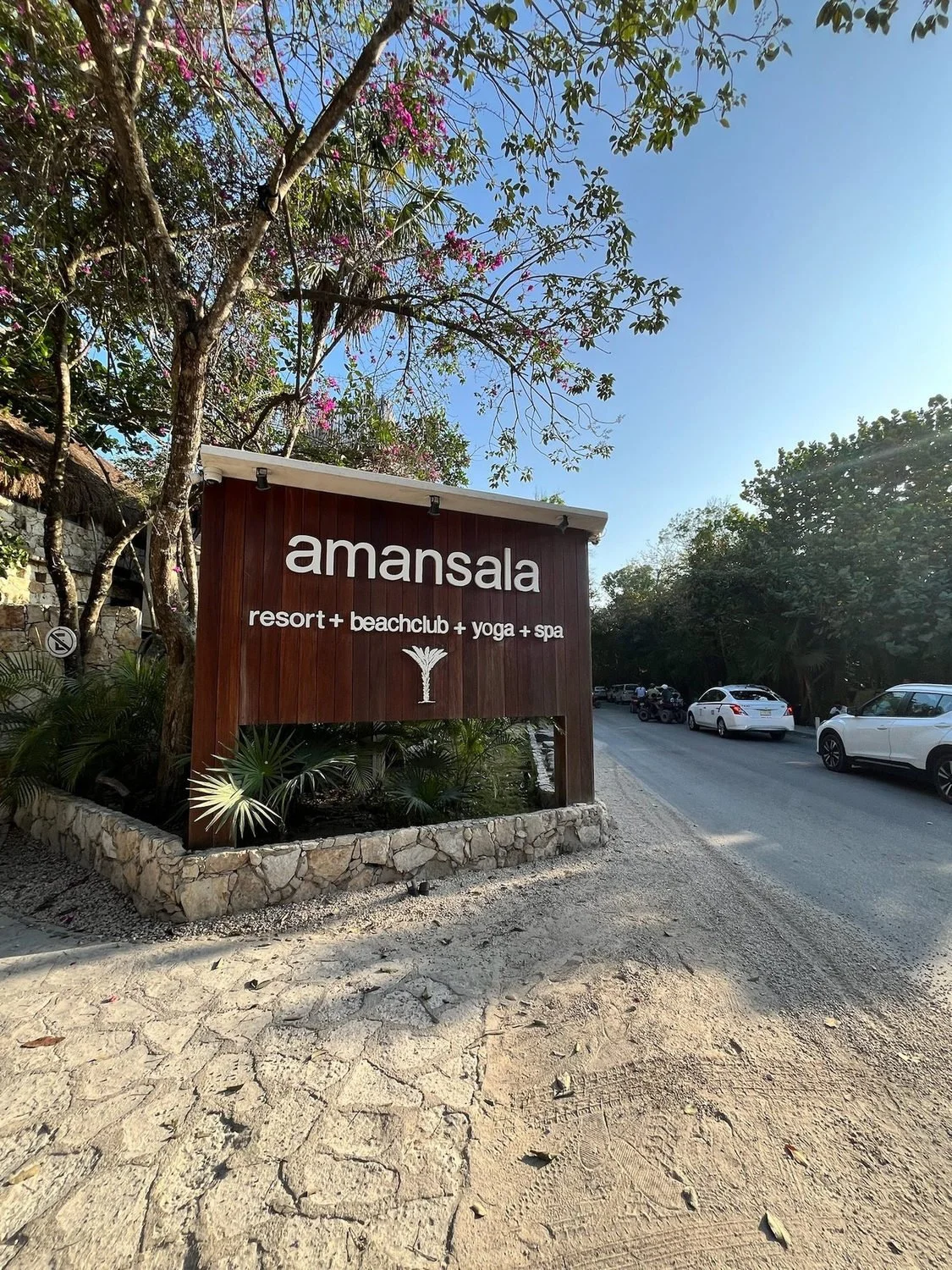 Amansala resort entrance sign beside a tree-lined road in Tulum.