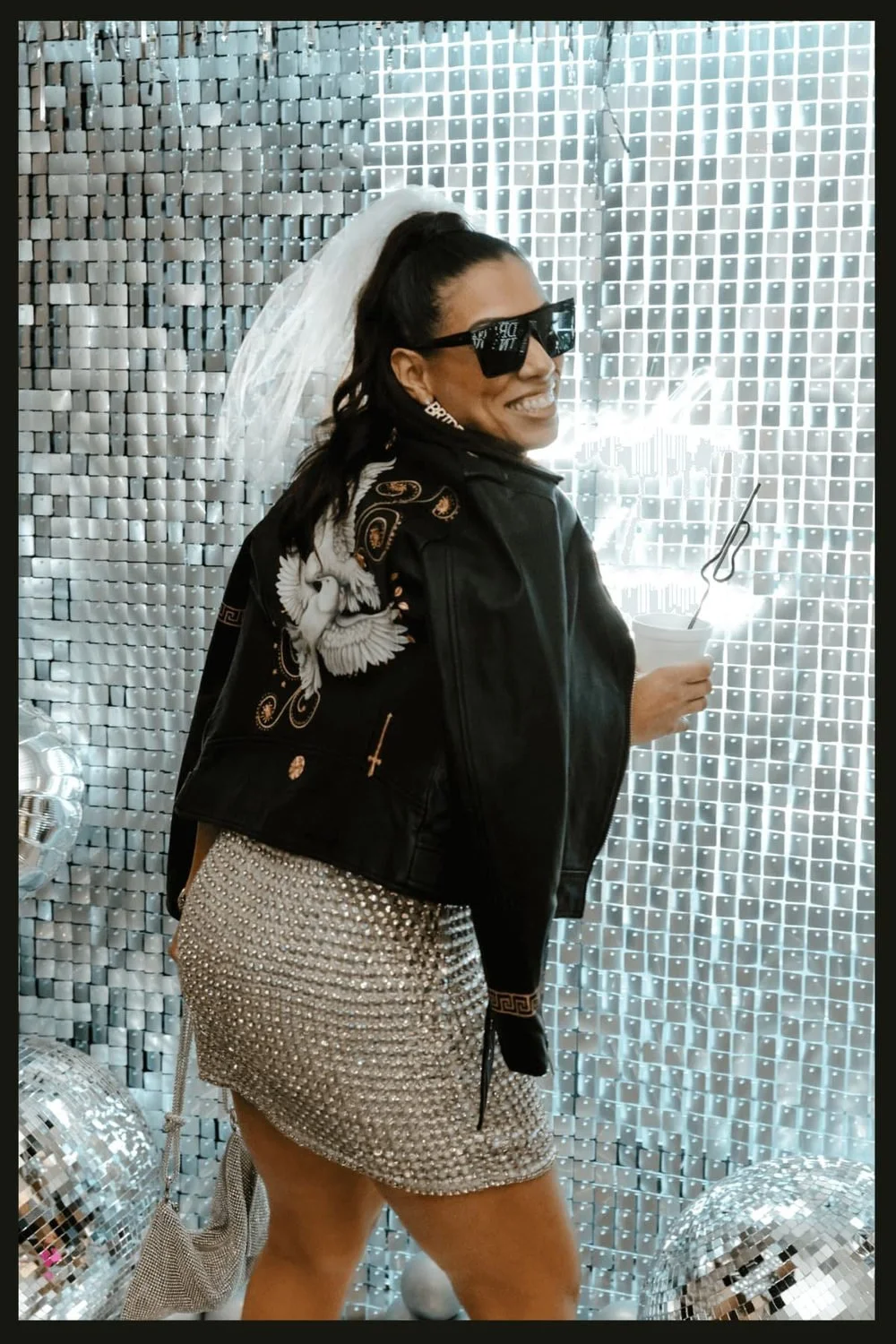 Bride wearing a veil, sunglasses, an embroidered black jacket and a silver skirt in front of a sequin backdrop.