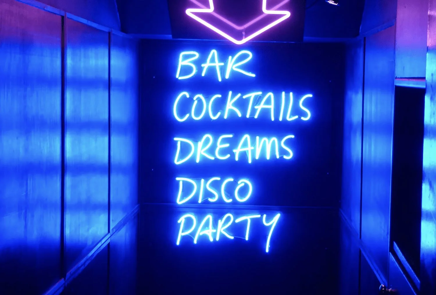 Blue neon sign reading Bar, Cocktails, Dreams, Disco, Party beneath a purple arrow.