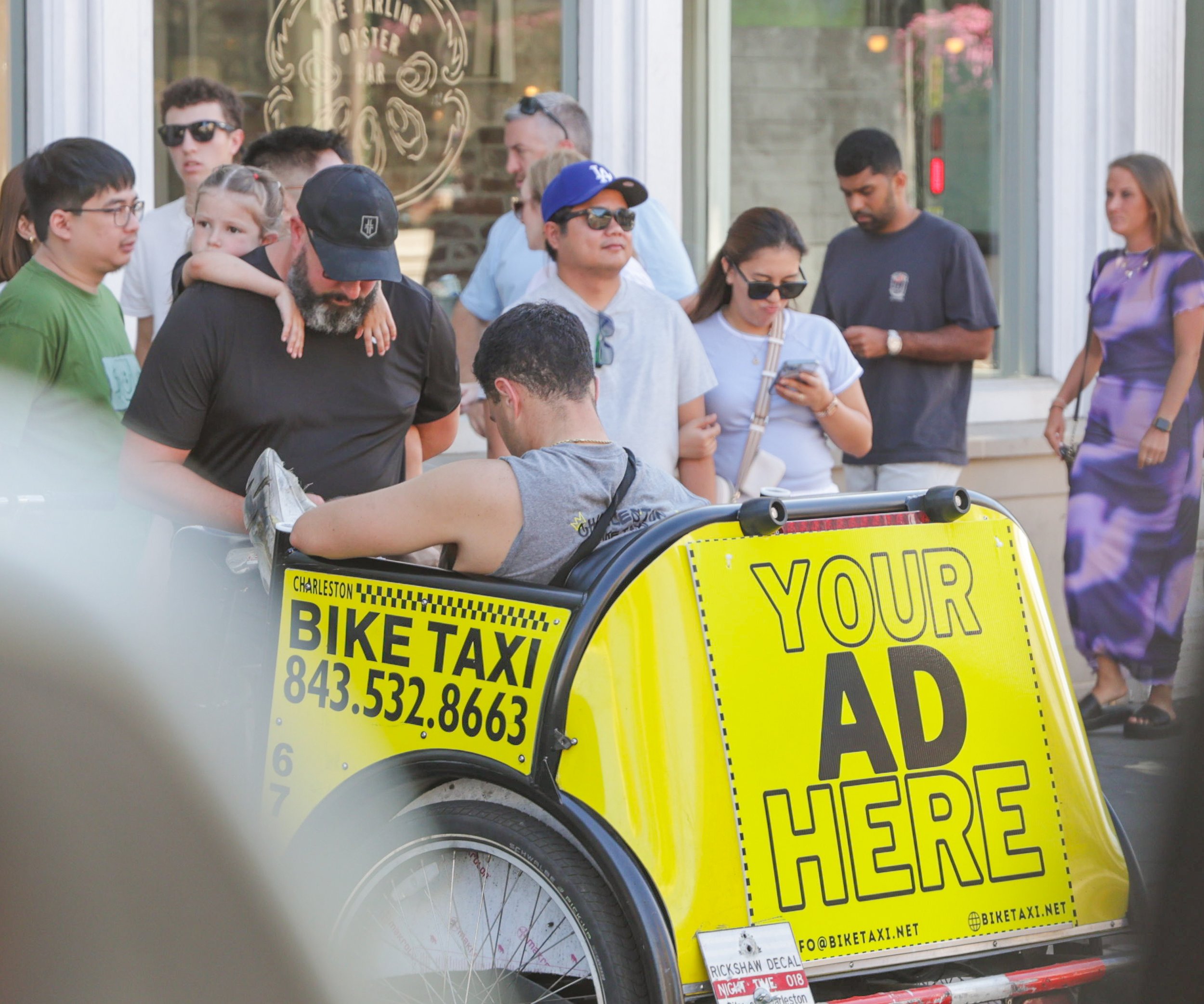 A man sitting in a yellow bike taxi with a sign that reads "Your Ad Here" is surrounded by a diverse group of people, some of whom are looking at their phones or talking, in front of a shop with a view of pedestrians walking by.