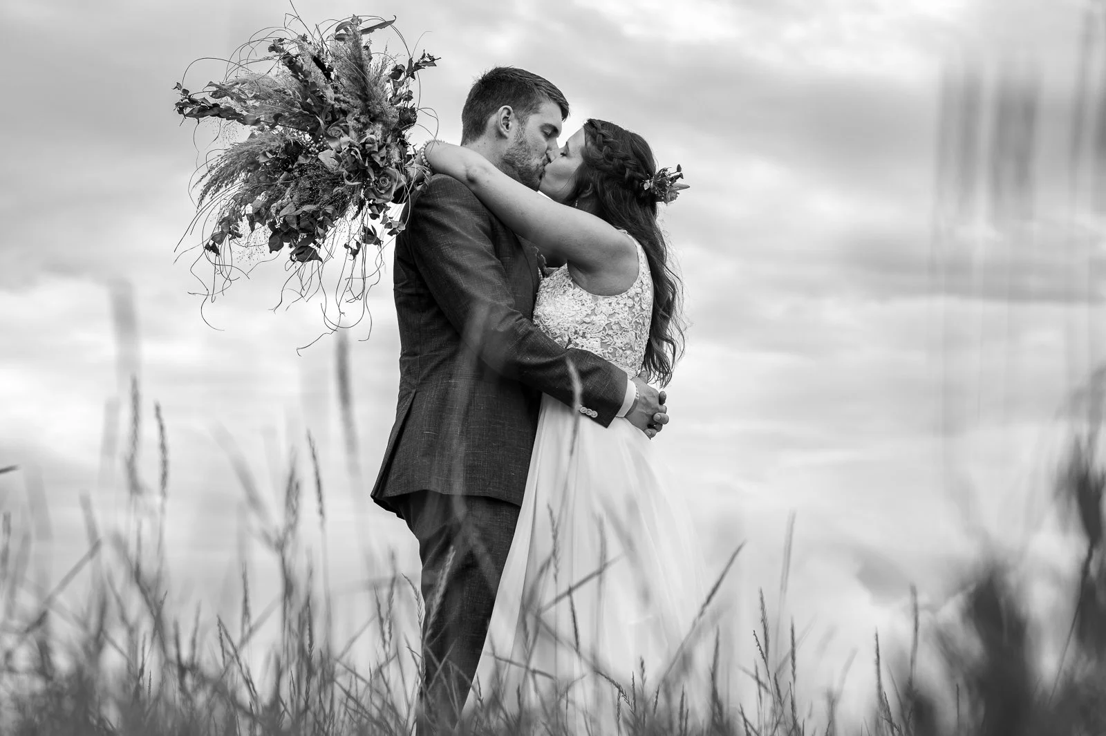 Ein Brautpaar küssend im Freien, schwarze und weiße Fotografie, die Frau hält einen Blumenstrauß, im Hintergrund Wolken und Wiese.